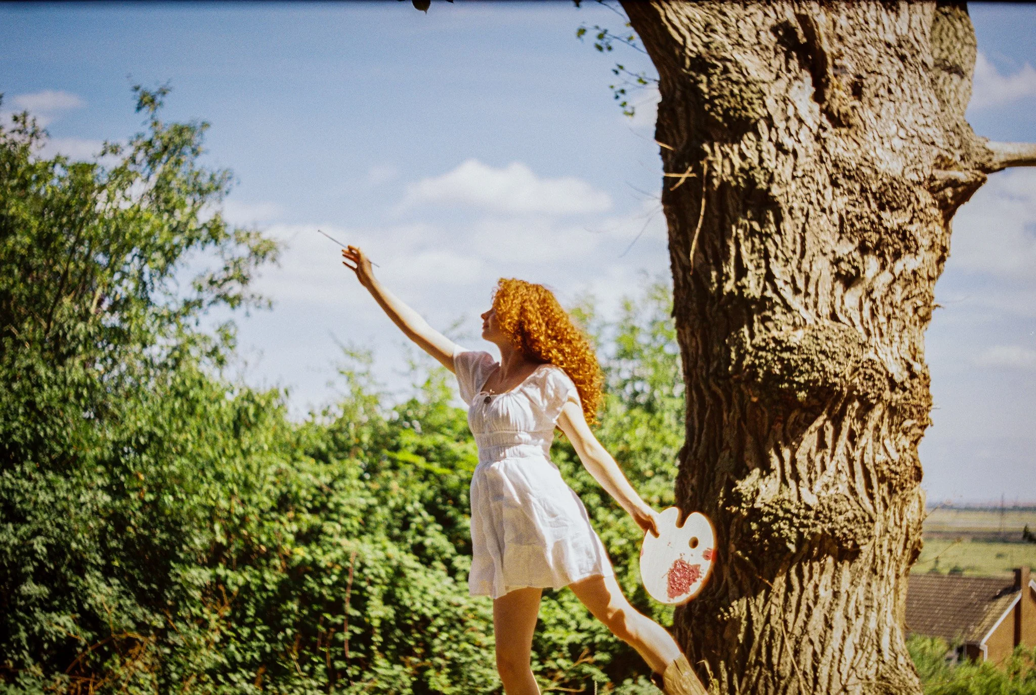 A woman with curly red hair wearing a white dress is dancing and spinning around outdoors, holding hands with a child. They are next to a large tree with a textured bark, and the background shows green bushes and a blue sky with some clouds.