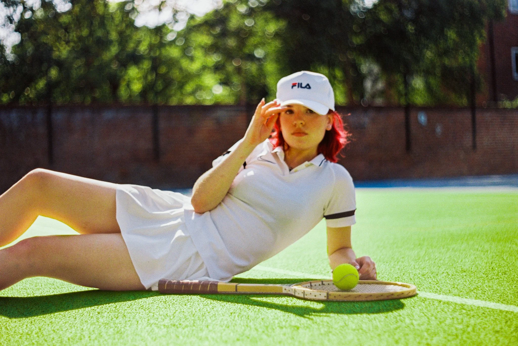 A woman with red hair wearing a white FILA cap and white outfit is lying on a green tennis court, holding a tennis ball near a tennis racket and tennis basket.