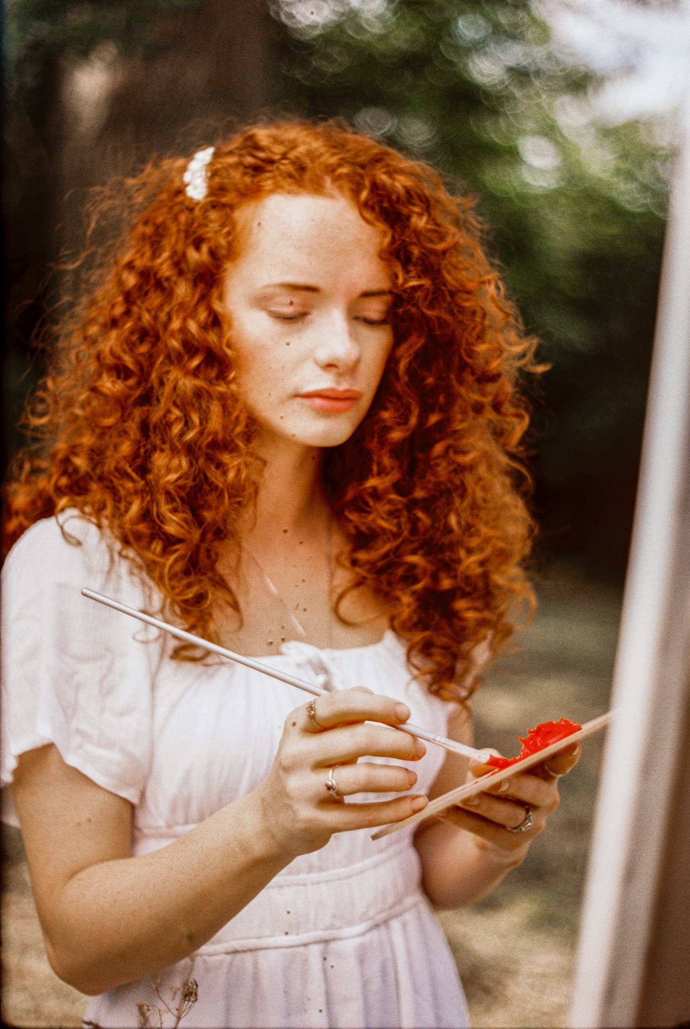 A young woman with curly red hair wearing a white dress and holding a paint palette and brush, with her eyes closed and a serene expression.