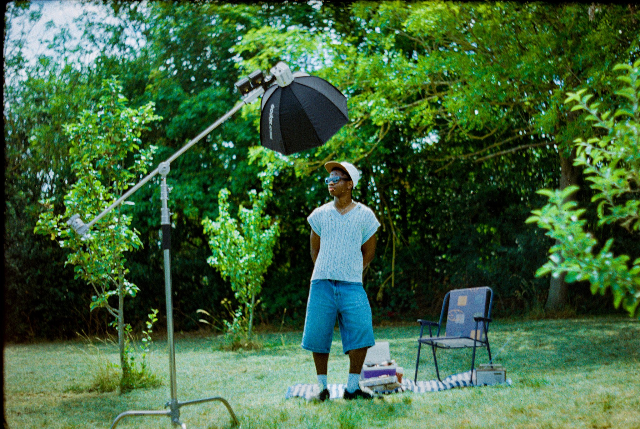 Person standing outdoors with an umbrella attached to a stand, surrounded by greenery, with chairs and equipment nearby.