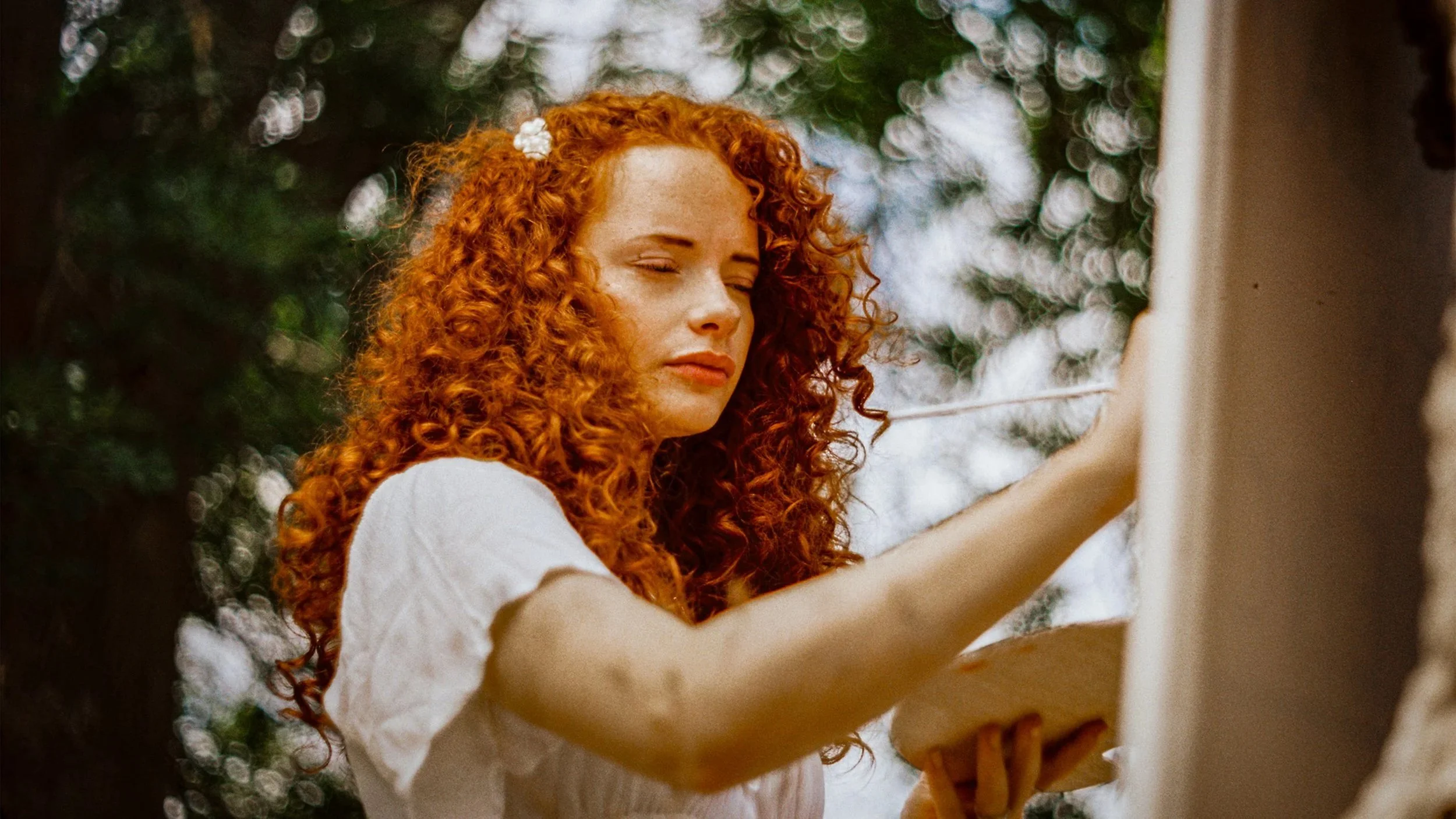 A young woman with curly red hair, wearing a white top, is planting a white flower in her hair outdoors under trees with a blurred background.