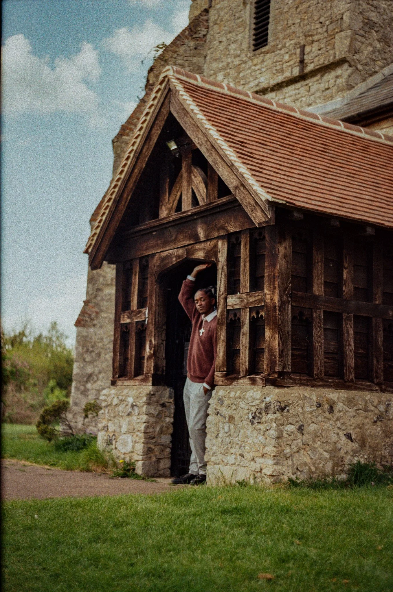 A man in a brown sweater and white pants standing in a wooden structure with stone walls, partially obscured by the wooden framework, outside a stone building.