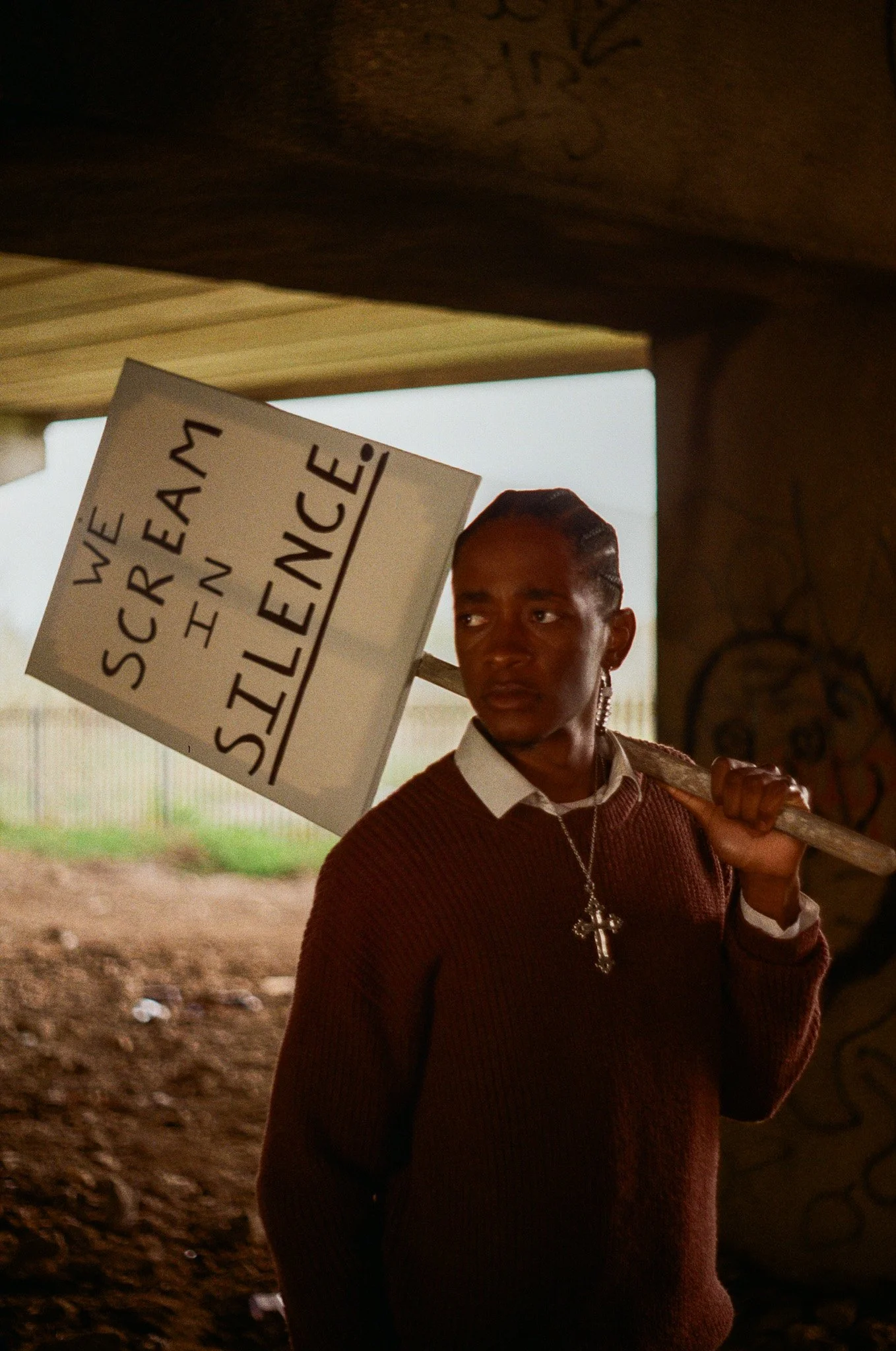 A woman with braided hair, wearing a sweater and shirt, holding a sign that says 'We are receiving testimonies,' standing in a shaded outdoor area with a stone wall and a person in the background.