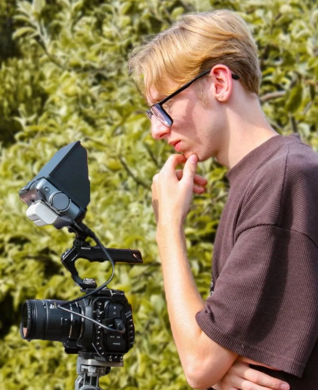 Sam Moore operating a professional video camera outdoors, with green foliage in the background.