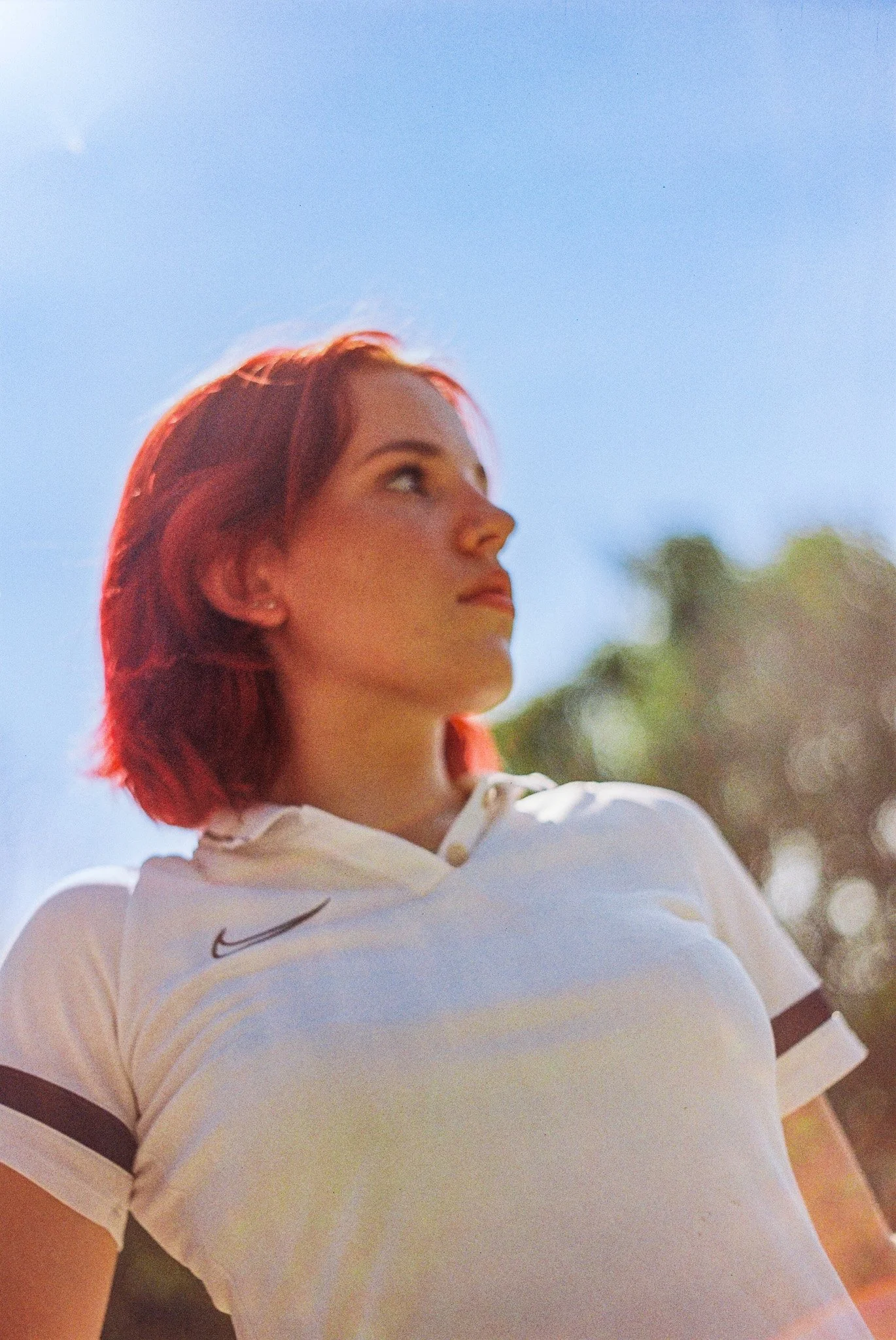 A woman with red hair wearing a white sports shirt with black stripes on the sleeves, standing outdoors against a blue sky with blurred trees in the background.
