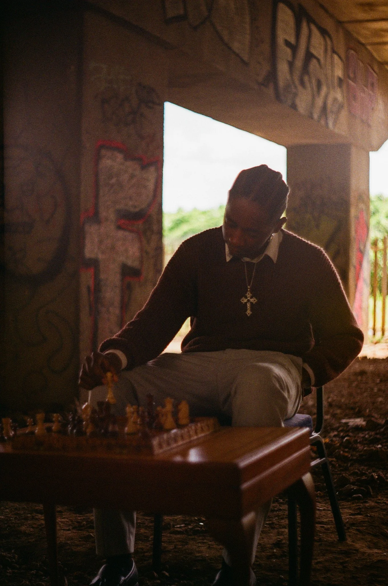 A man wearing glasses and a cross necklace is sitting on a chair indoors, with sunlight coming through an opening behind him. There is graffiti on the walls around him and a chessboard on a table in front of him.