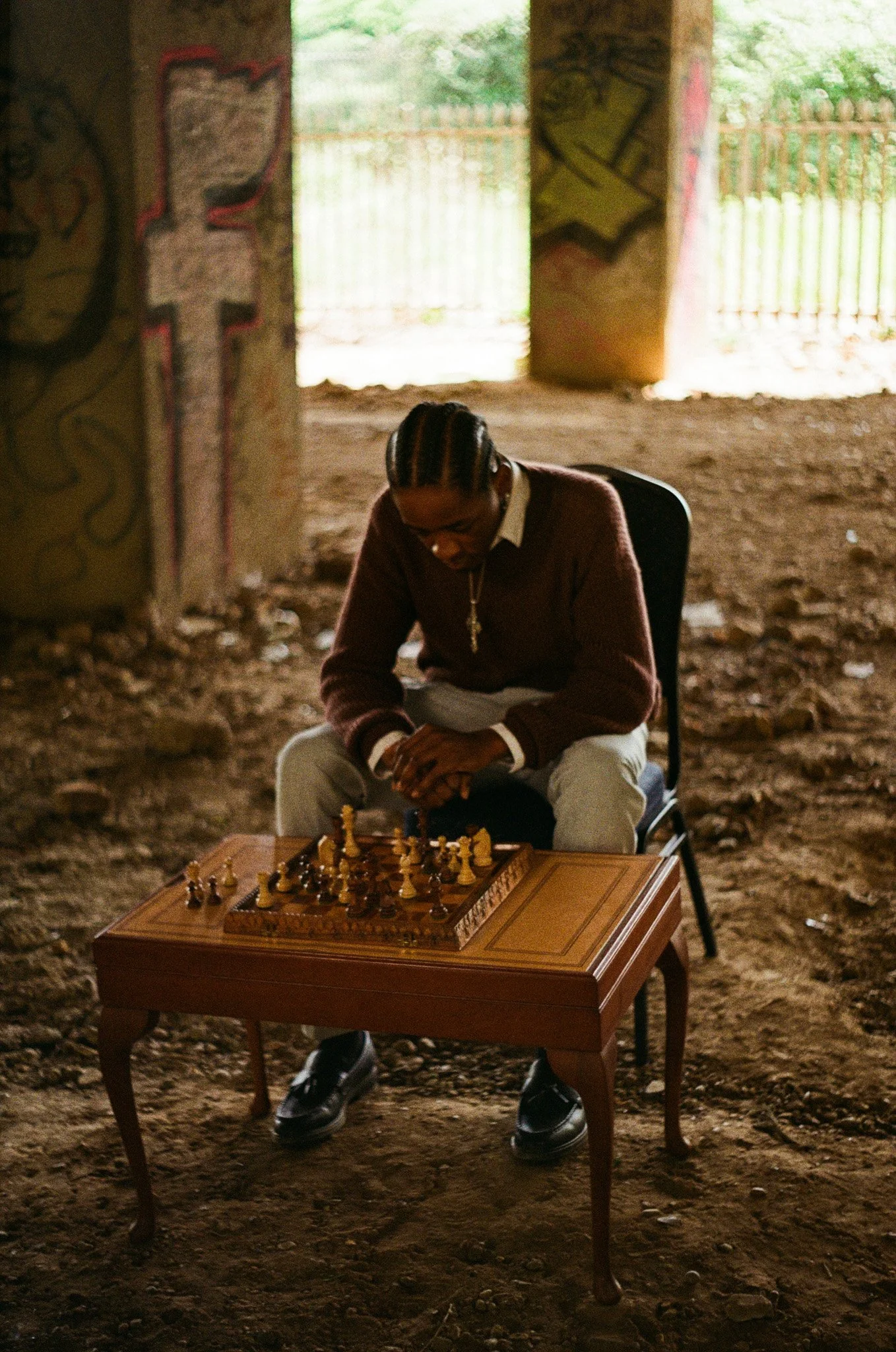 A person with braided hair sitting and playing chess at a small wooden table in an underground or covered area with graffiti on the wall and a dirt floor.