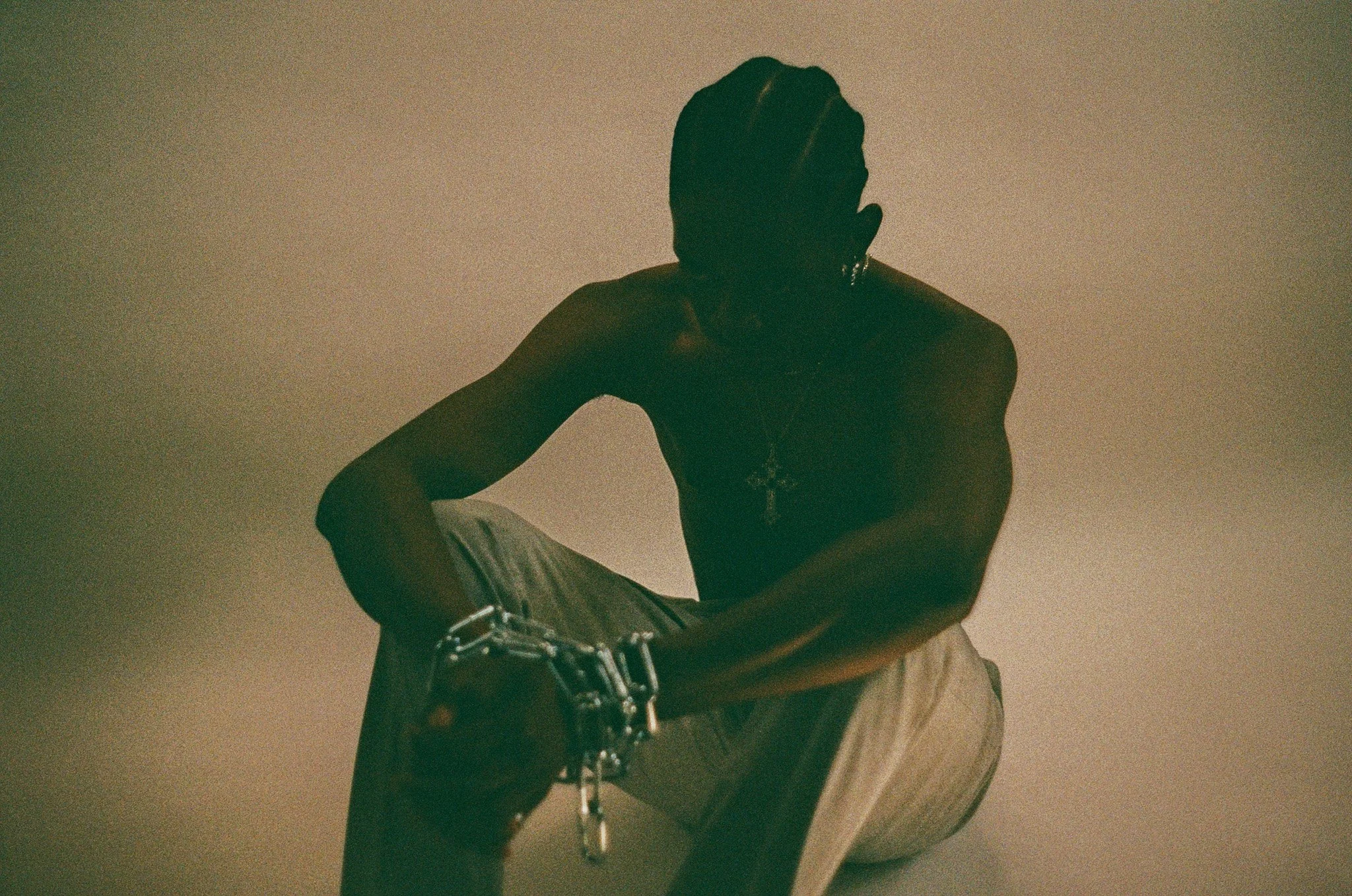 A person with dark skin sitting on a wooden surface, wearing a cross necklace and a black shirt, with their face obscured and hand covering their face.