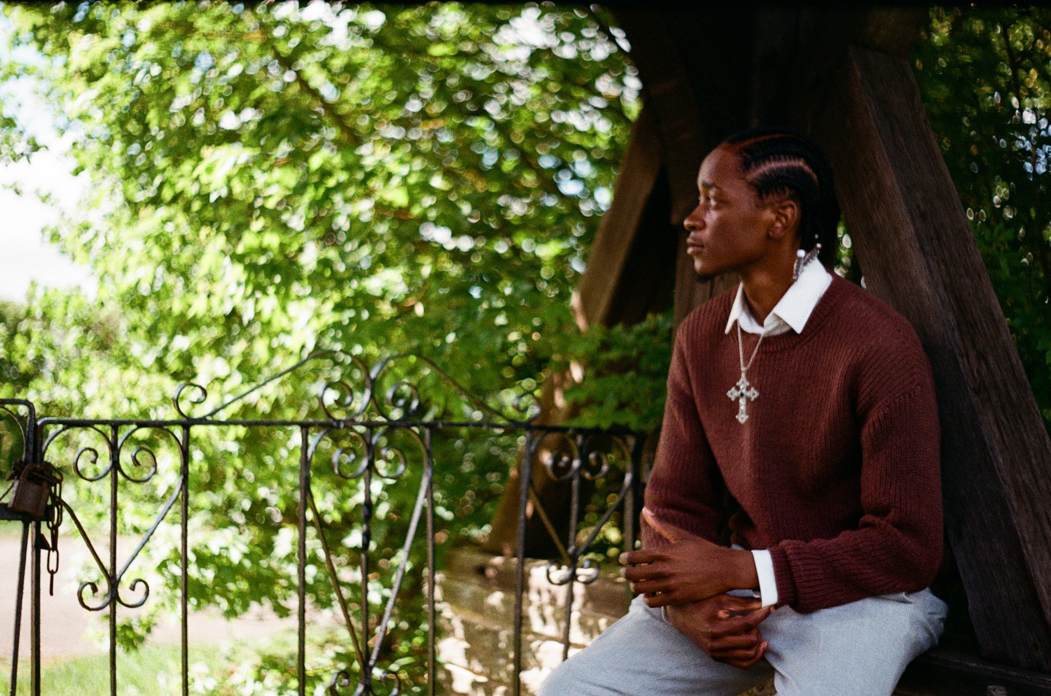 A woman with braided hair, wearing a cross necklace, white shirt, and brown sweater, sitting outside under a tree with green leaves, near a black wrought iron fence.
