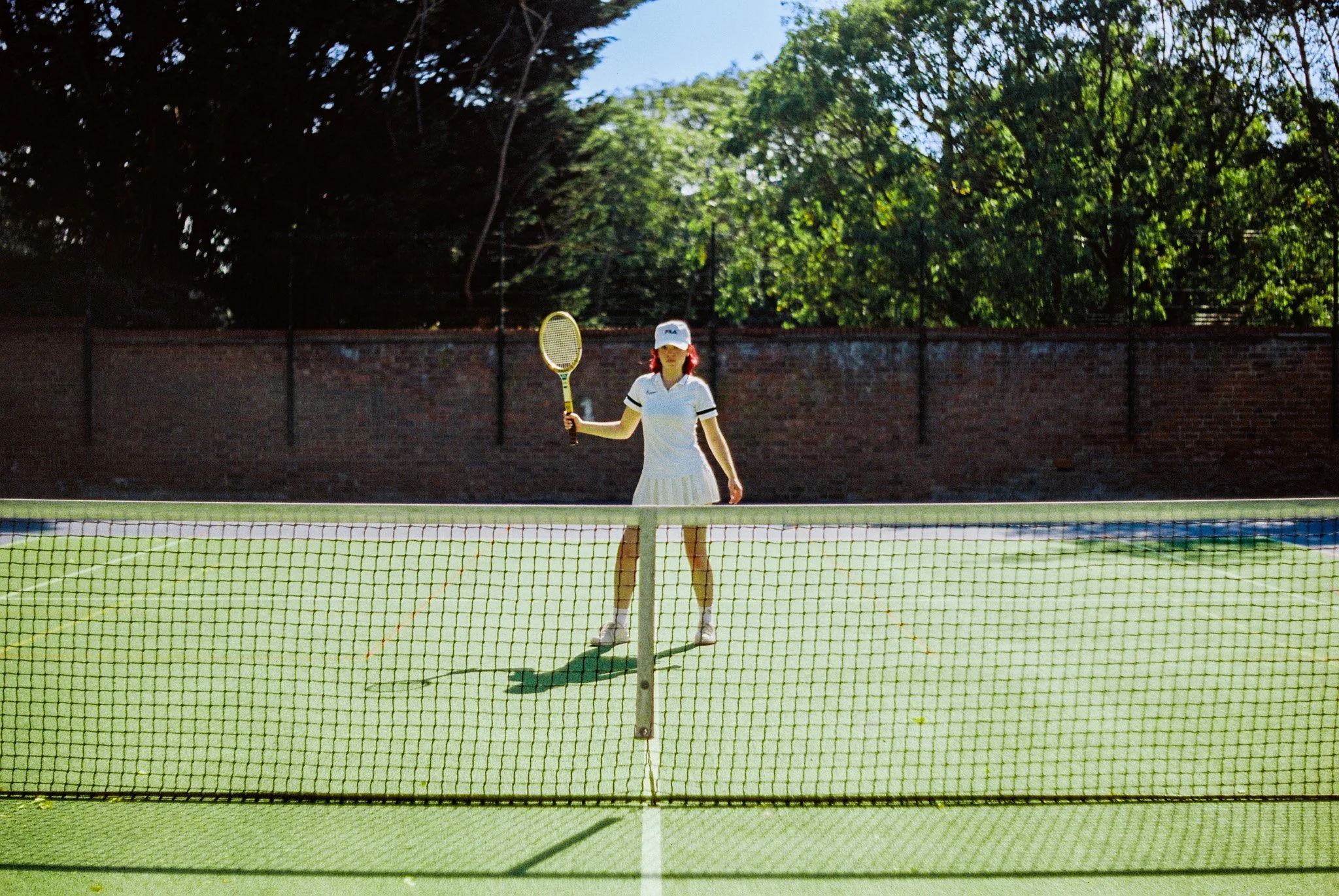 A woman stands on a tennis court holding a tennis racquet, wearing a white hat and tennis outfit, with a brick wall and trees in the background.