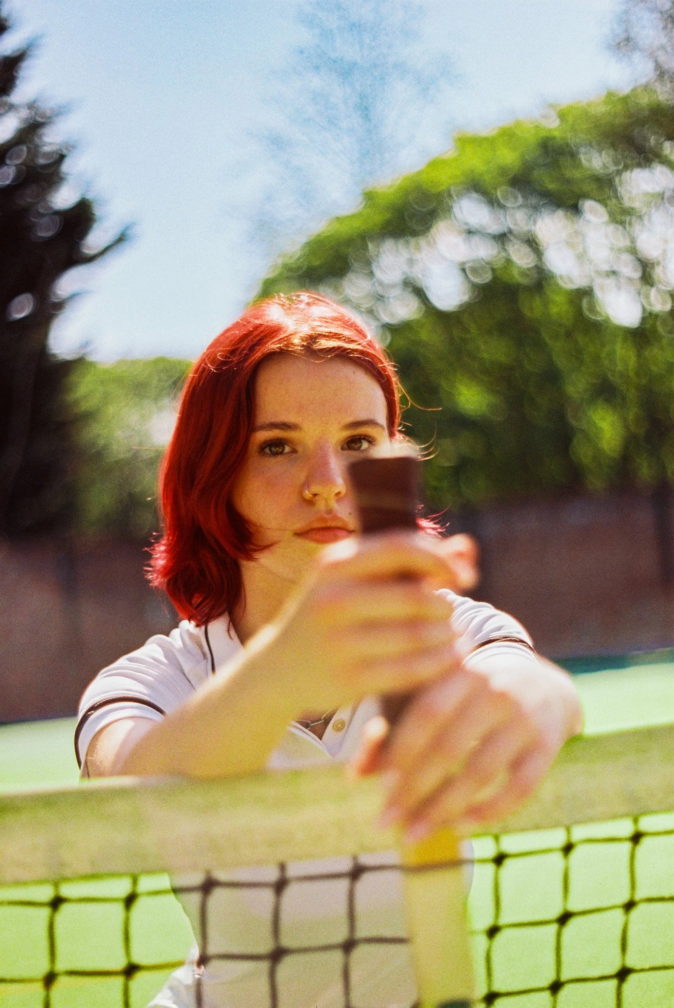 A young woman with red hair taking a selfie through a tennis net on an outdoor court, with trees and a blue sky in the background.