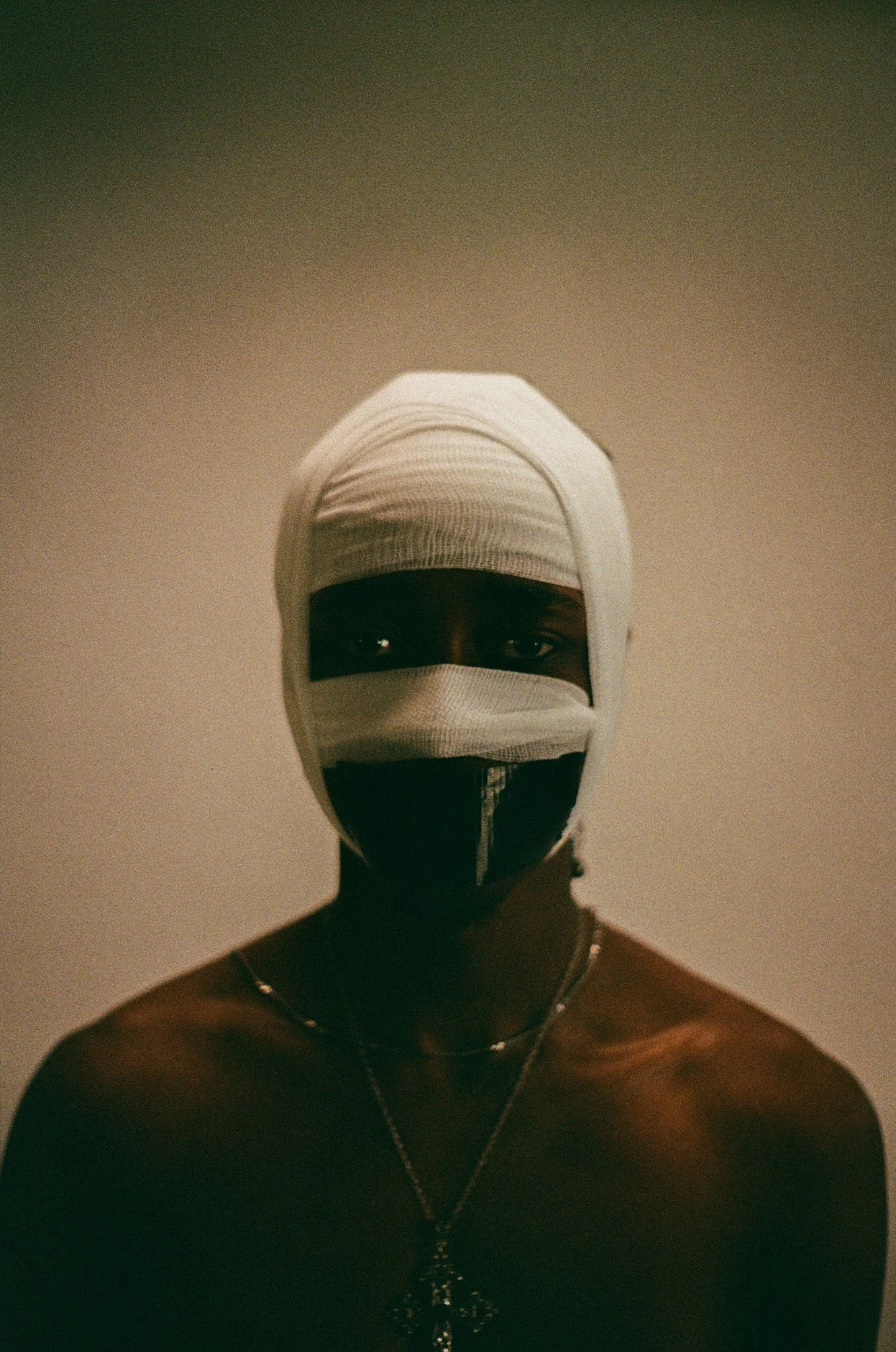 A person with dark skin wearing a white bandage wrap over their head and face, with black eye masks and a zipper across the face, is shirtless and wearing layered necklaces, standing against a neutral background.