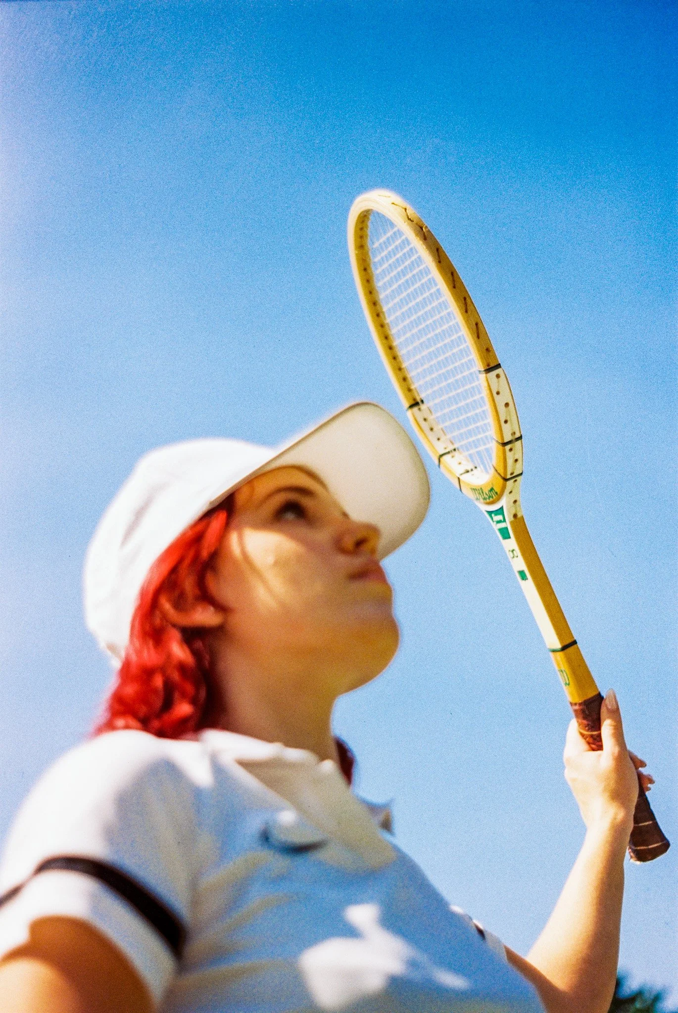 A woman with red hair wearing a white polo shirt and a white cap, holding a tennis racket, looking up against a clear blue sky.
