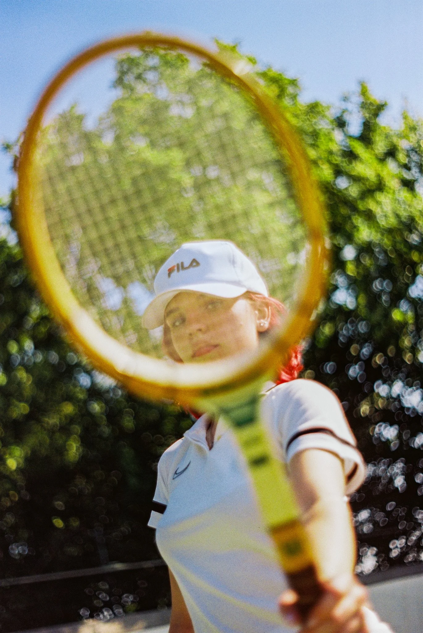 A tennis player holding a tennis racket in front of the camera, with trees and blue sky in the background.