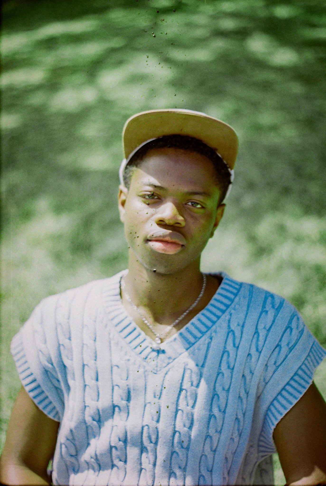 A young man wearing a beige baseball cap and a light blue T-shirt with text, standing outdoors with a blurred green background.