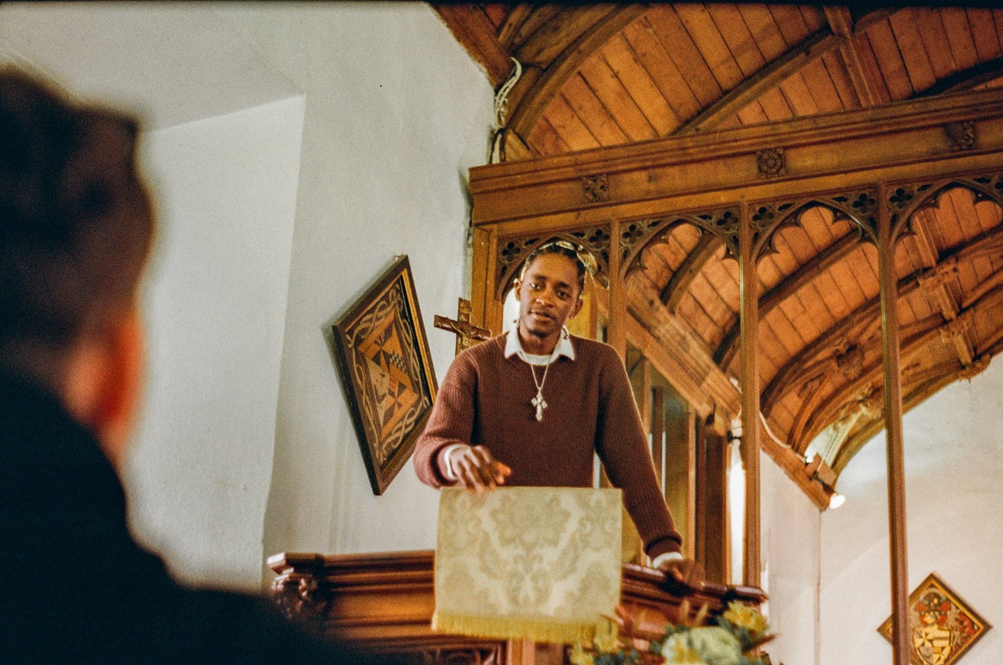 A young man with dark hair and dress shirt, wearing a cross necklace, is standing behind a pulpit in a church, speaking or gesturing towards a congregation. The church features wooden arches and religious artwork.