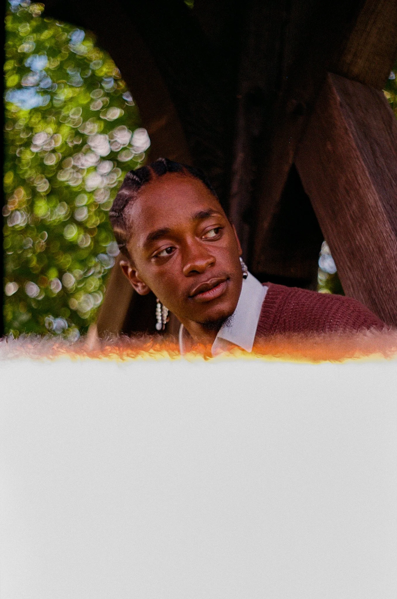 A person with braided hair wearing earrings and a maroon sweater, looking to the side, outdoors with green foliage in the background.