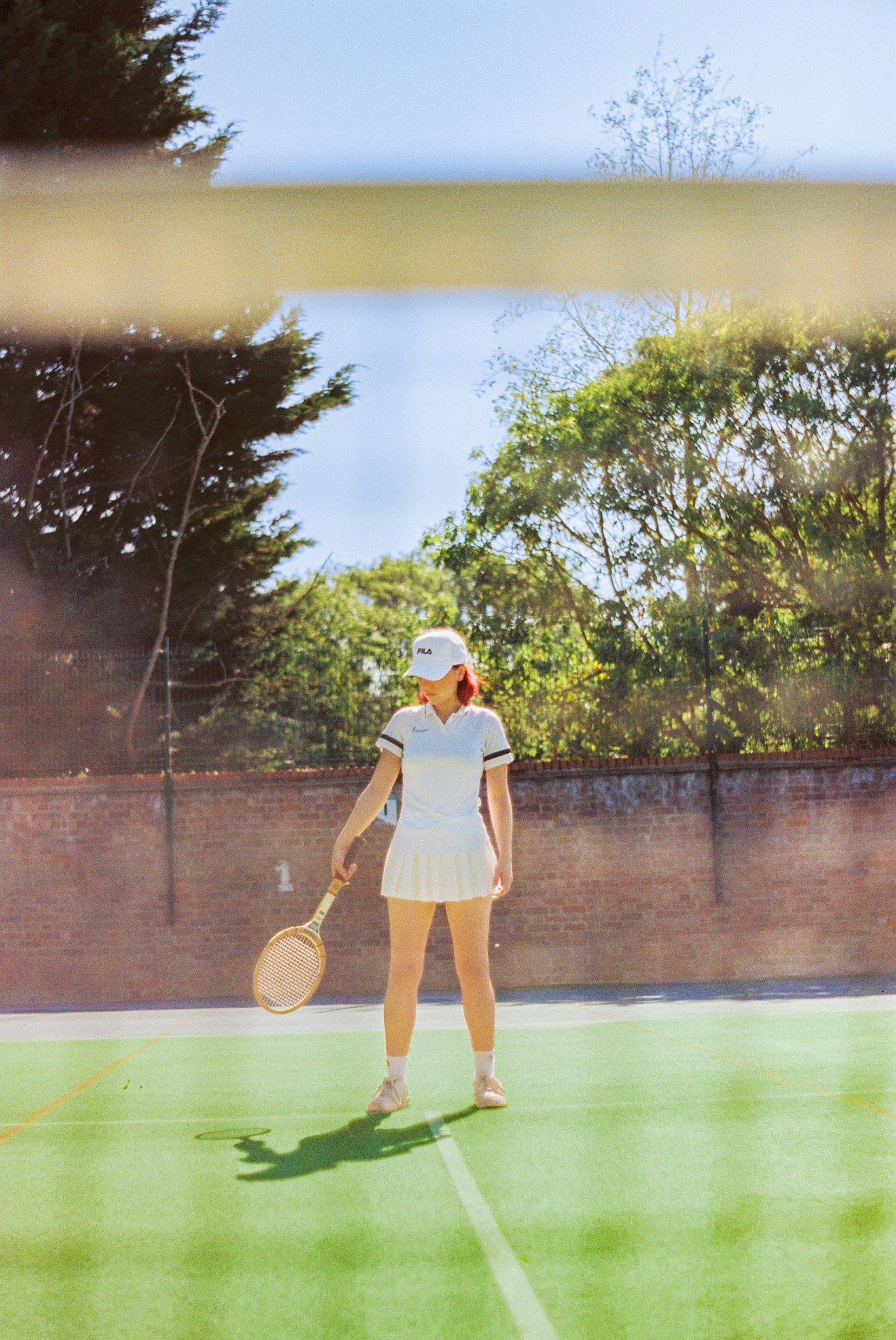 A young woman in tennis attire standing on a tennis court holding a tennis racket, with a brick wall and trees in the background.