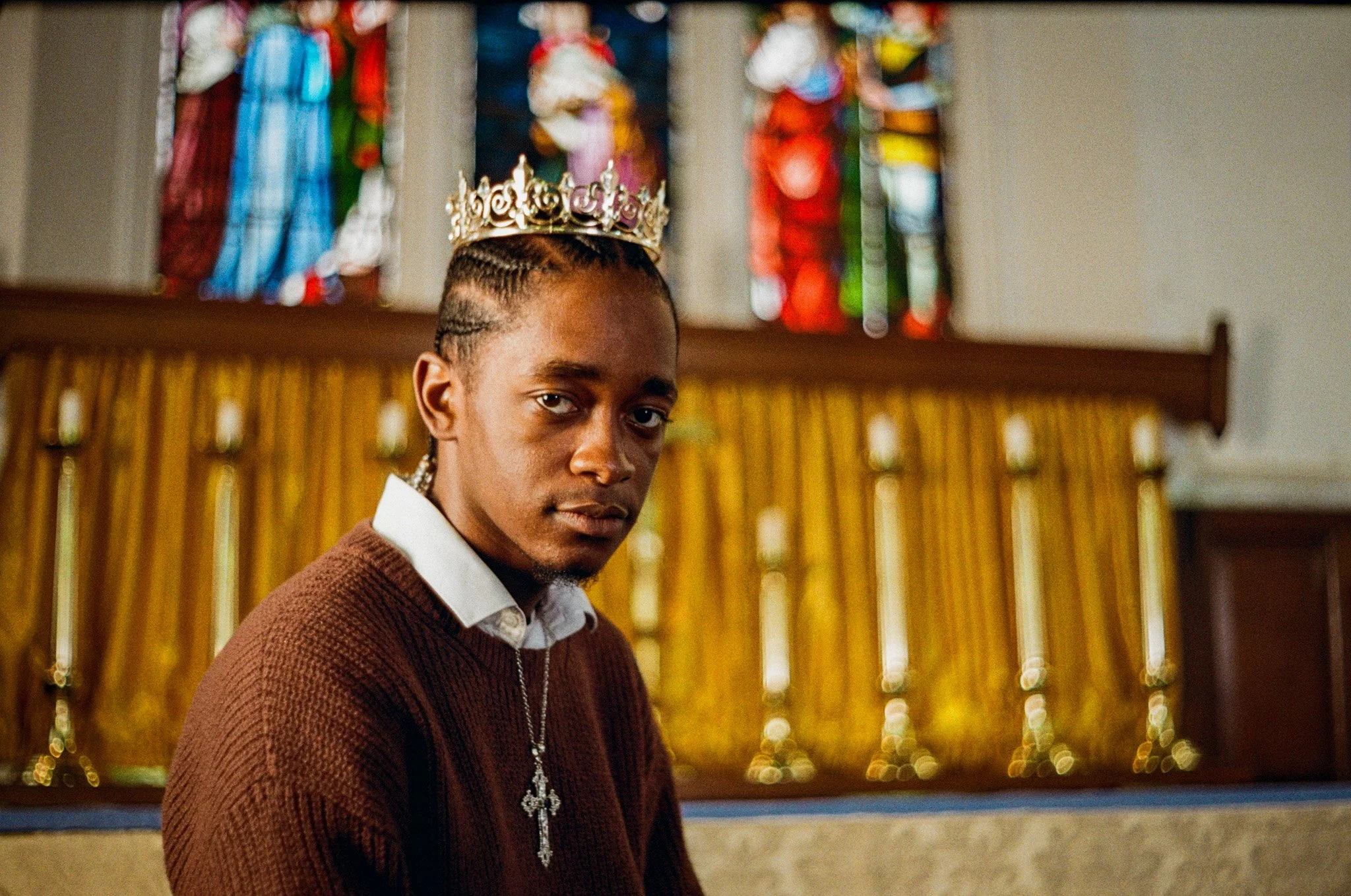 Young man wearing a crown with the word 'birthday' on it, standing indoors with colorful stained glass windows in the background.