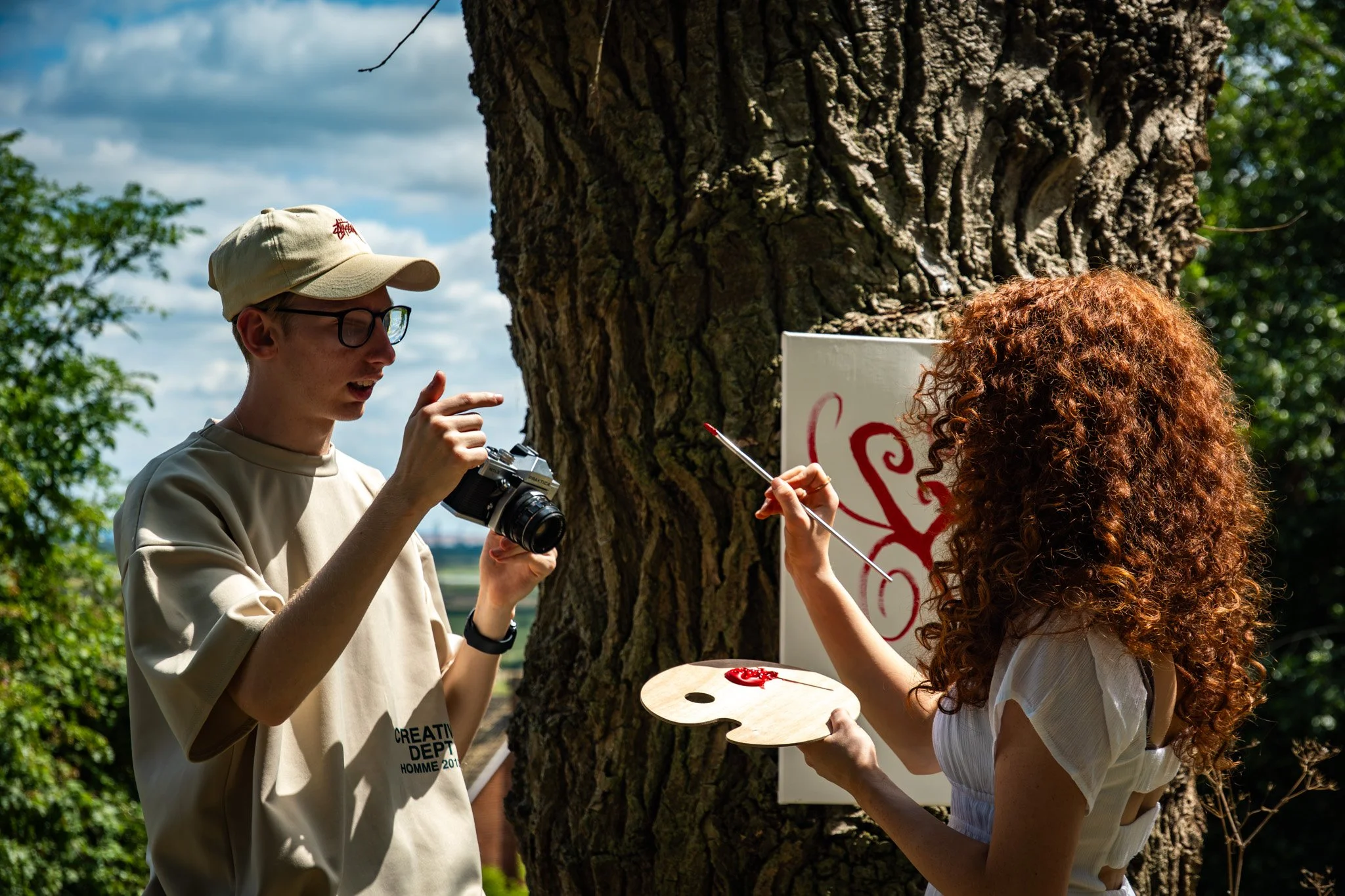 Sam Moore directs an actor near a large tree, the actor holds a paint palette with red paint and a brush, with a partially visible painting that has red lettering is seen in the background.