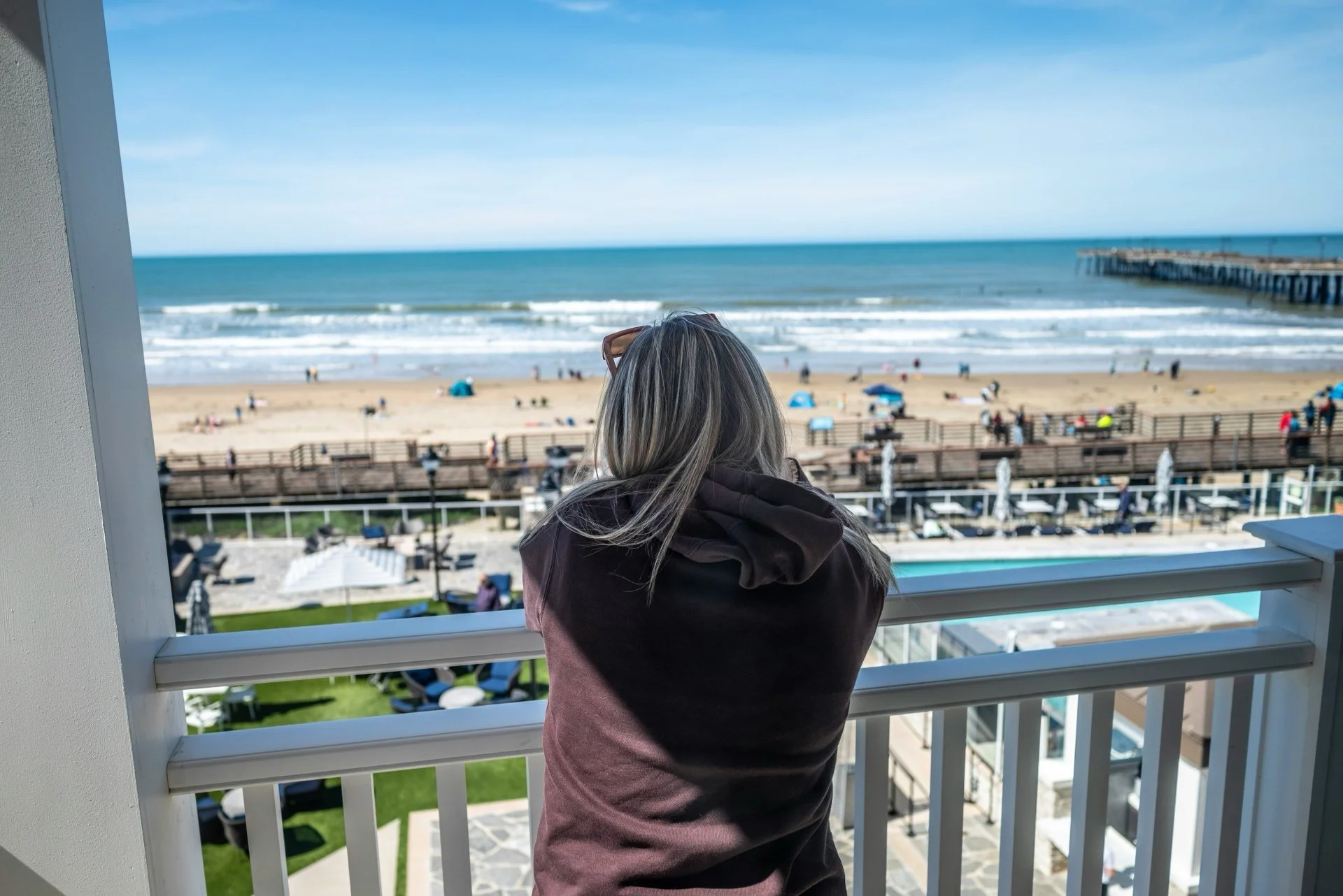 A person with gray hair and a pink hoodie stands on a balcony overlooking a sandy beach, ocean waves, and a pier in the distance with a clear blue sky.