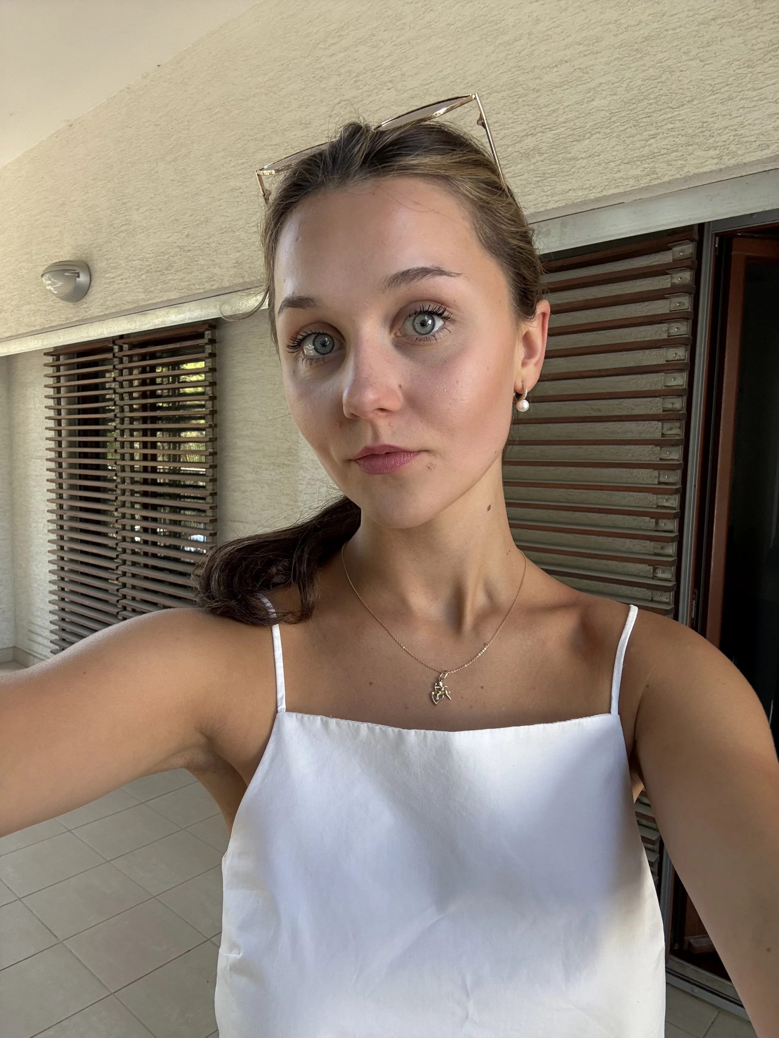 A young woman with blue eyes and light brown hair taking a selfie outdoors. She is wearing a white spaghetti strap top, pearl earrings, and a necklace with a small pendant. She has sunglasses resting on her head and is standing in front of a wall with horizontal wooden slats and a window.
