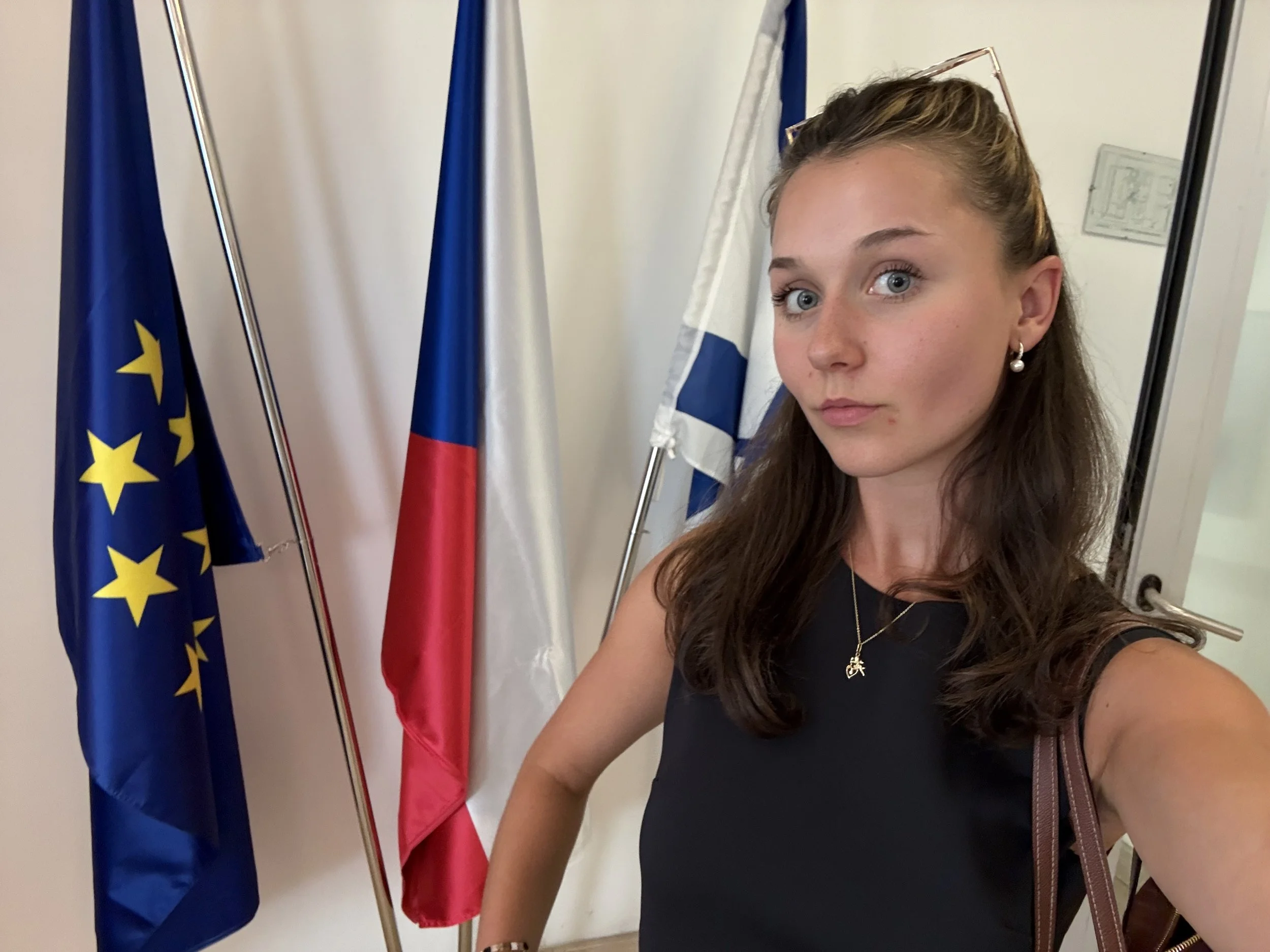 A young woman with brown hair, wearing earrings and a necklace, is taking a selfie in front of three flags: European Union, France, and Israel. She is wearing a dark top and has a brown strap bag.