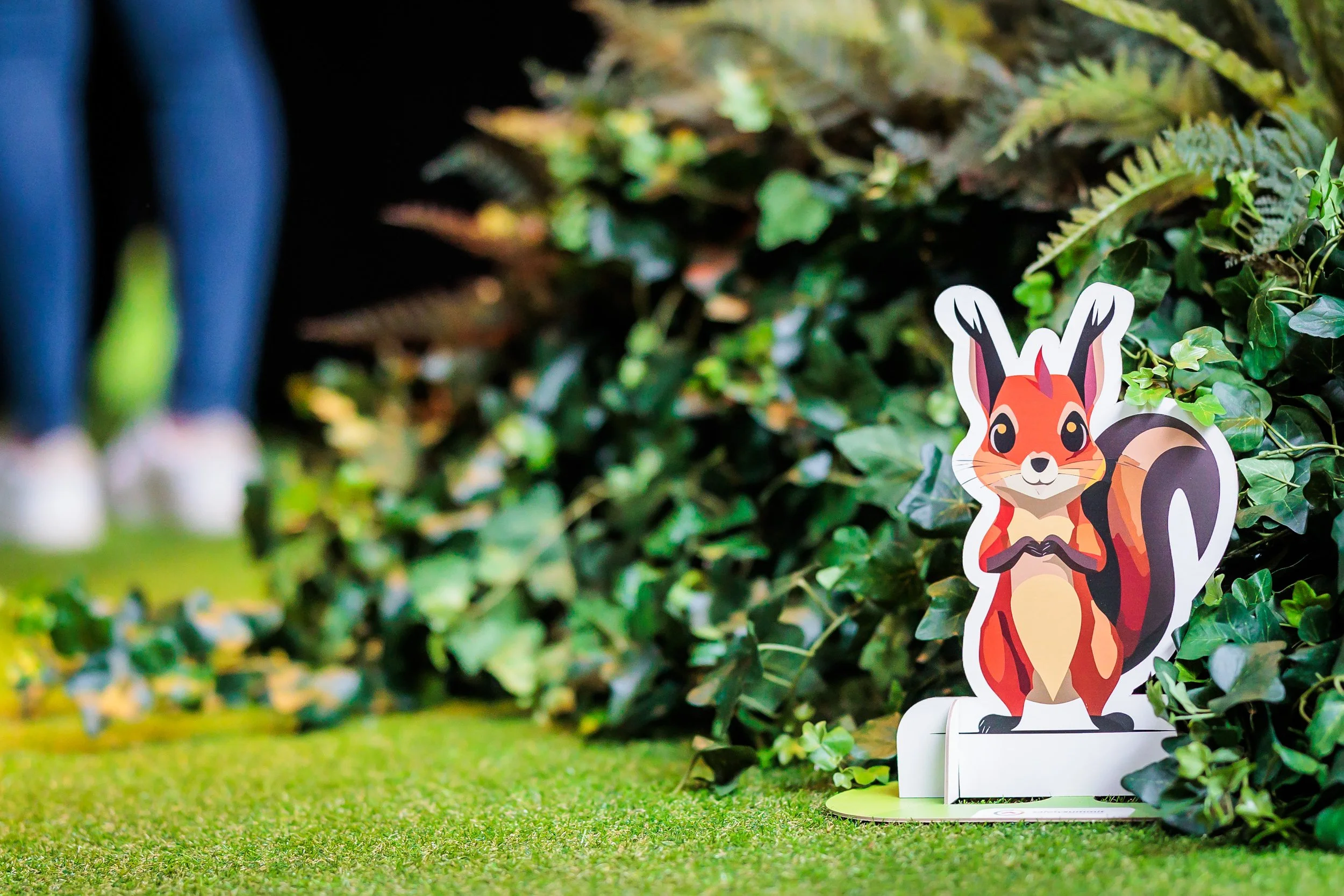 A cartoon deer cutout is positioned in front of a green leafy hedge, with a blurred person standing behind in blue jeans. The ground is covered with green grass.
