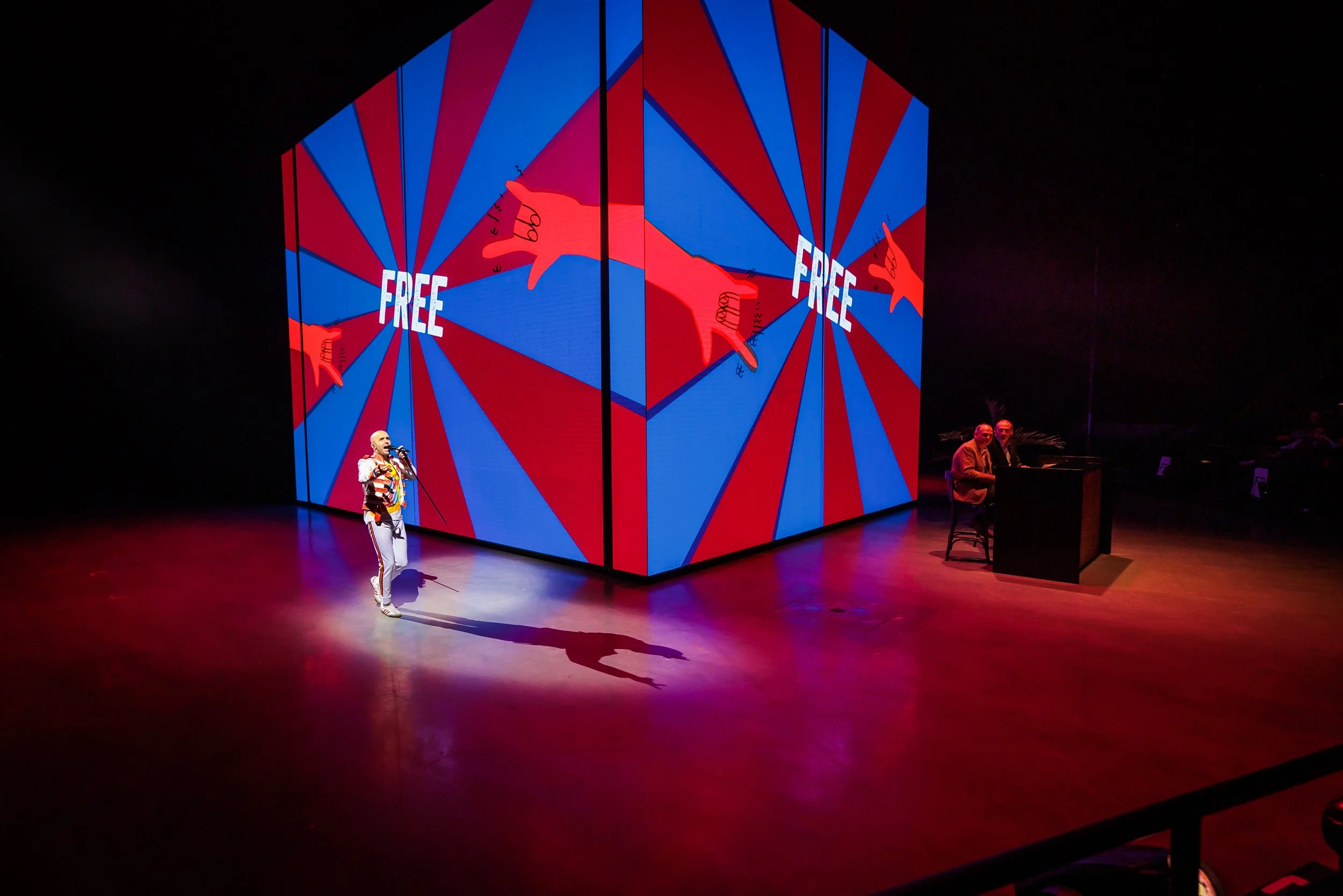 Performer singing on stage with a large, colorful, geometric backdrop displaying the word 'FREE' and red hands pointing outward.