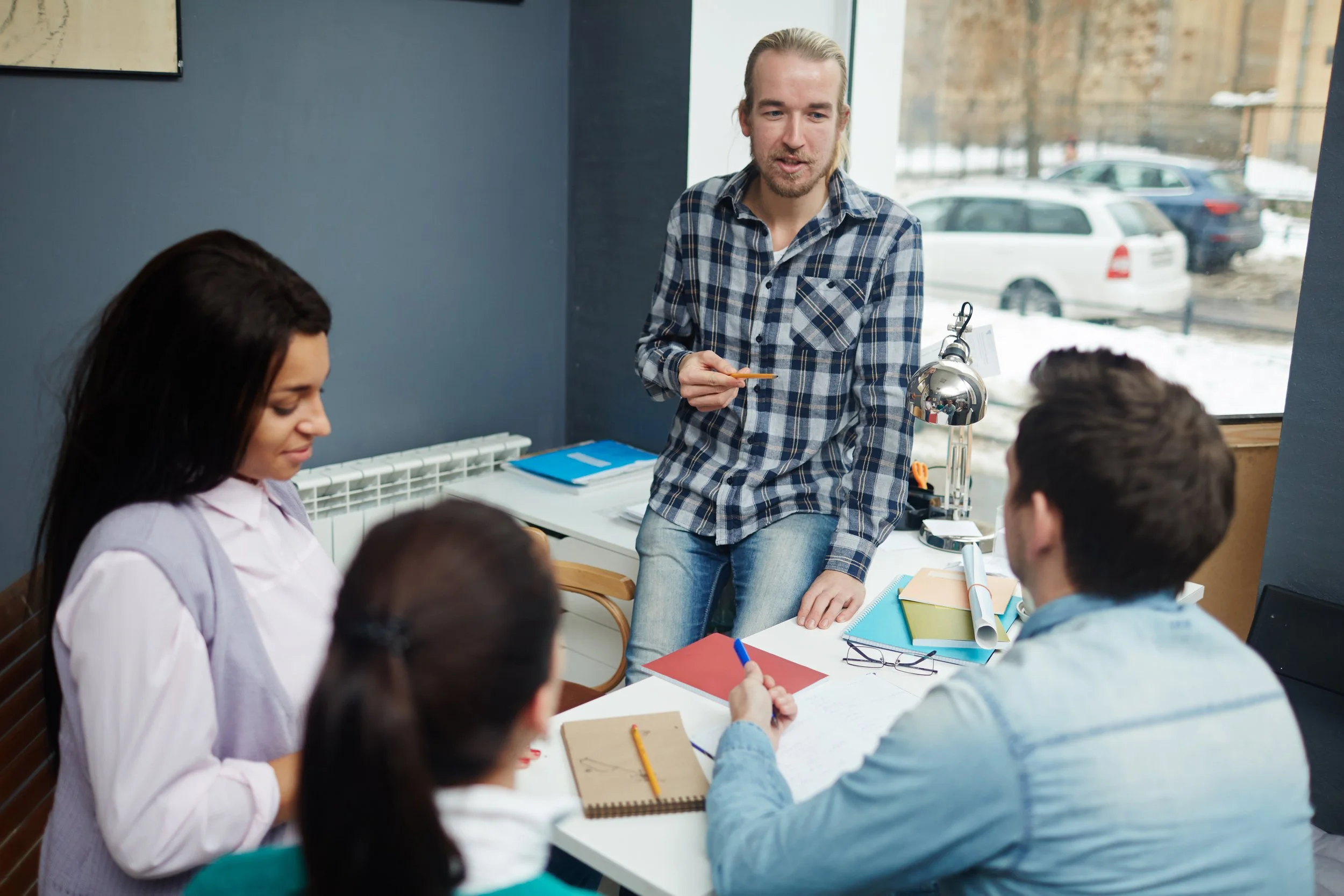 A man with long blonde hair and a beard, wearing a blue plaid shirt, is standing and talking to a group of people in a meeting room with large windows showing a snowy parking lot outside. The group includes women and men sitting at a table with notebooks, papers, and colored folders.