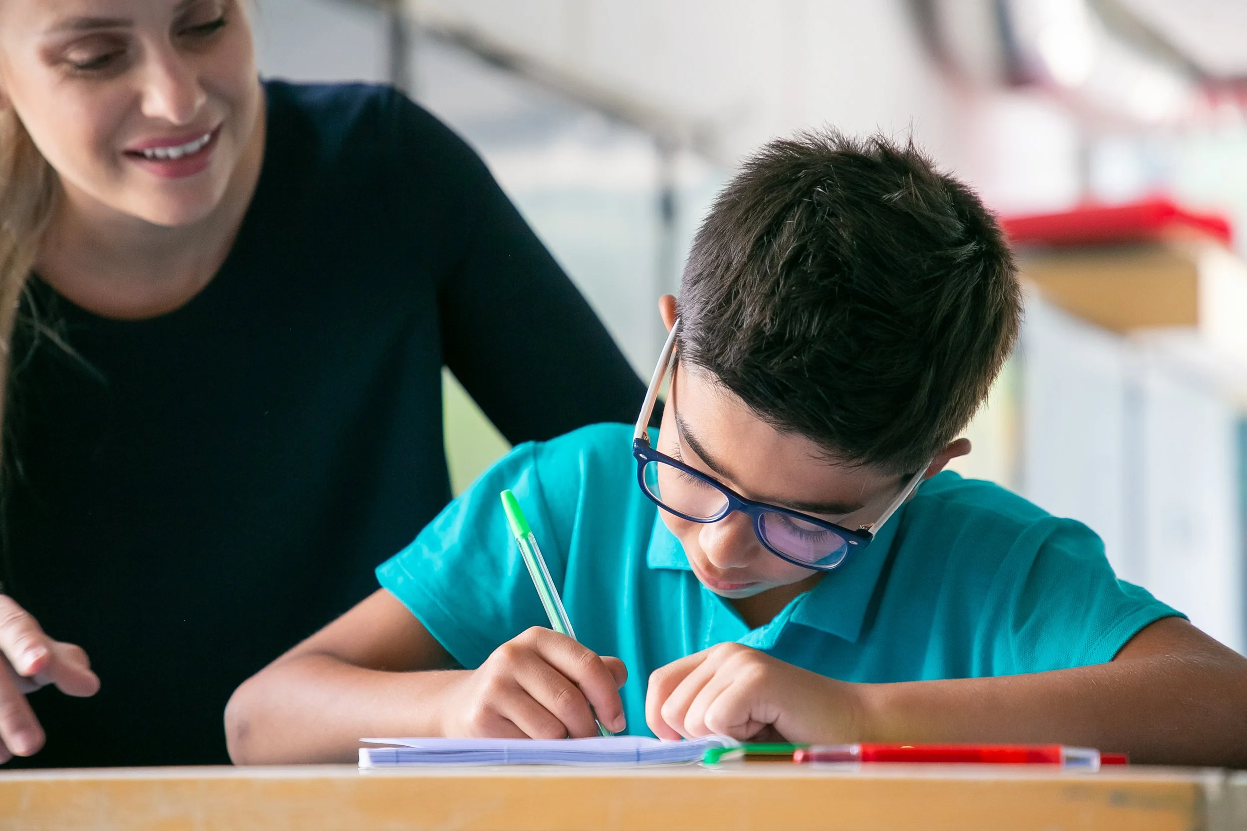 A teacher or adult helping a young boy with glasses write or do schoolwork at a desk in a classroom.
