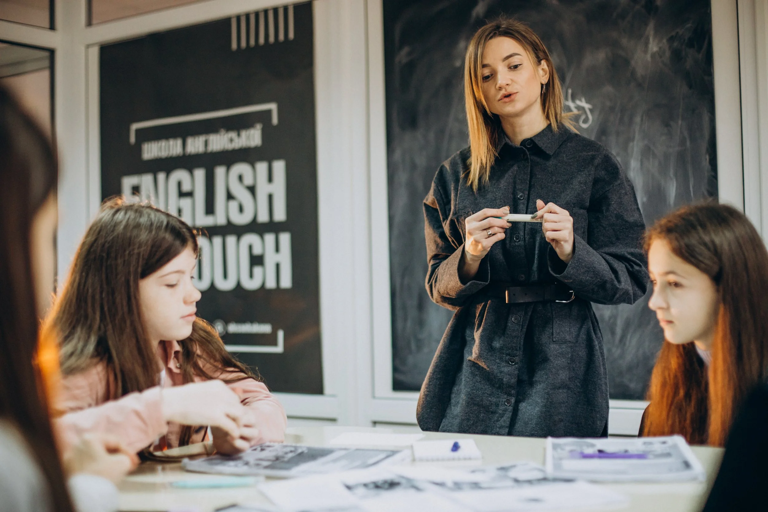 A teacher standing in front of a black chalkboard in a classroom with students seated at a table, some writing or drawing on papers, and a large sign on the wall behind reading 'English'.