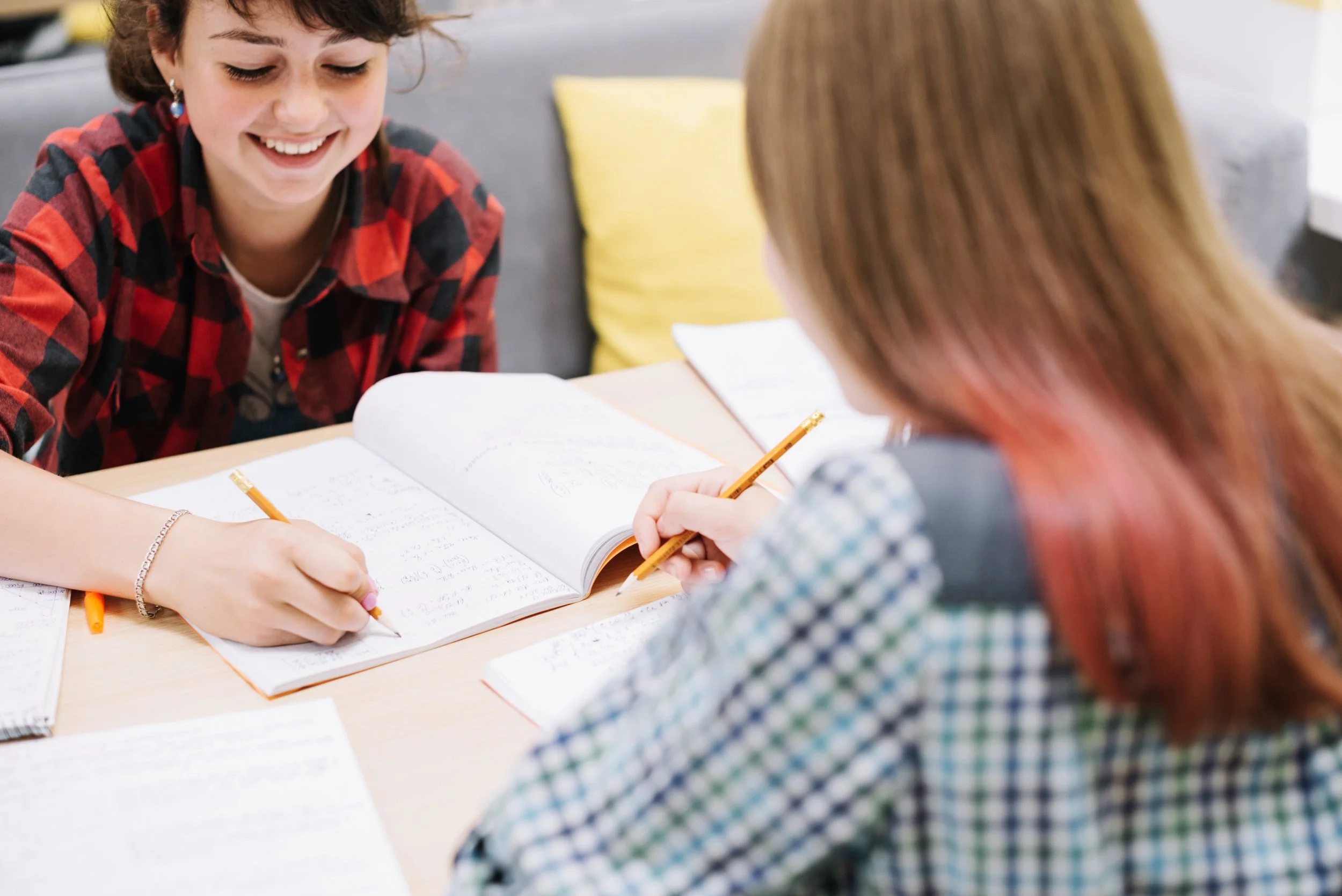 Two women sitting at a table with notebooks, tomatoes, pencils, and papers, engaged in a conversation or study session in a casual setting.