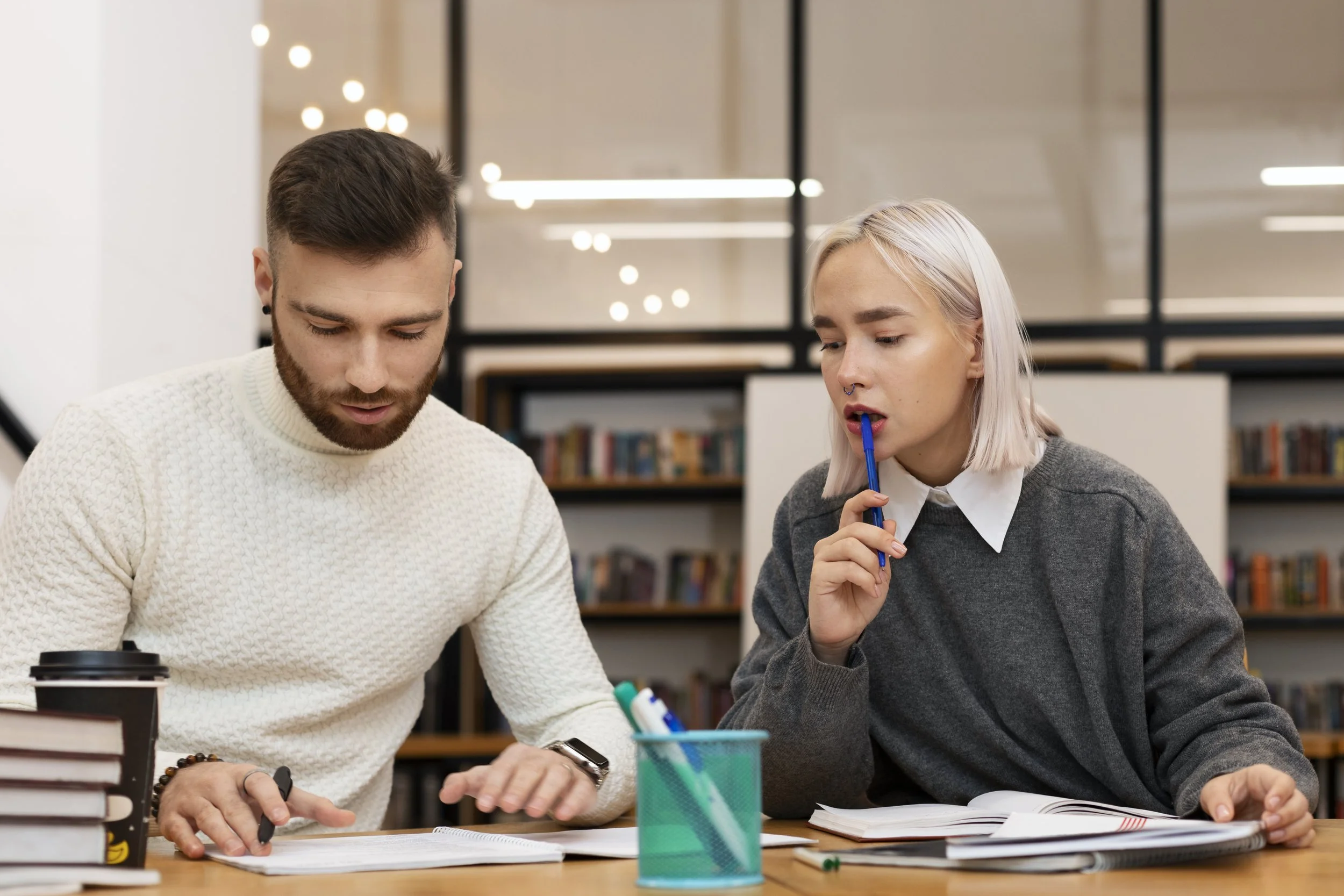 Two young adults, a man with dark hair and a beard and a woman with platinum blonde hair, sit at a table in a library or study area, engaged in reading or studying. There are books, notebooks, and writing supplies on the table, and a reusable coffee cup is visible on the left. The background features bookshelves and modern lighting fixtures.