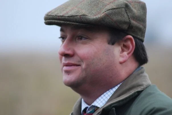 A man wearing a brown flat cap, a green jacket, and a collared shirt, standing outdoors with a blurred natural background.