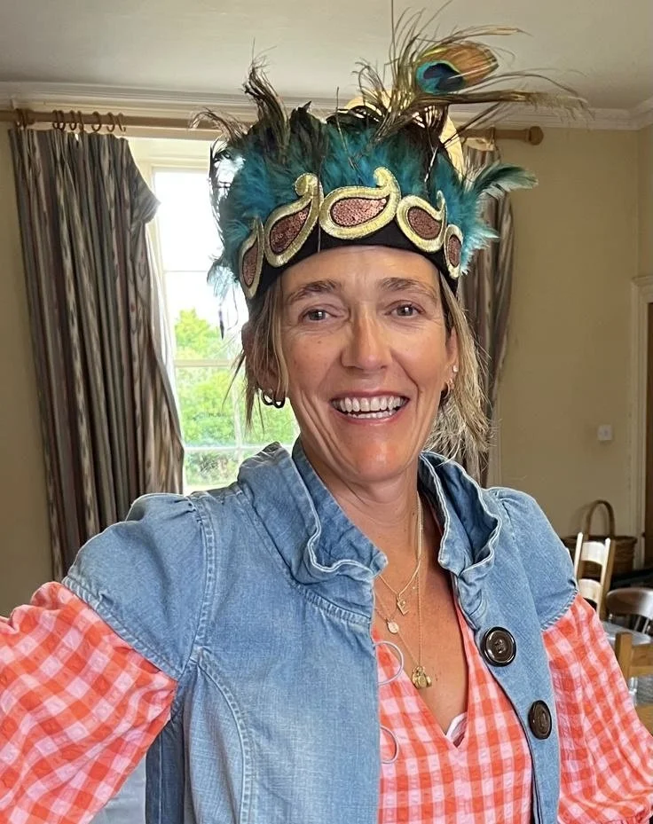 Woman wearing a colorful feathered headdress, a denim jacket, and smiling indoors.