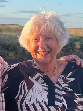 Smiling elderly woman with curly white hair outdoors, wearing a patterned shirt, during sunset.