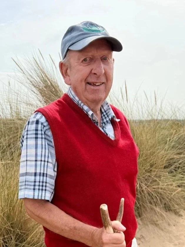 An elderly man wearing a gray cap, red vest, and checkered shirt, smiling and holding a walking stick while standing in front of tall grass at the beach.