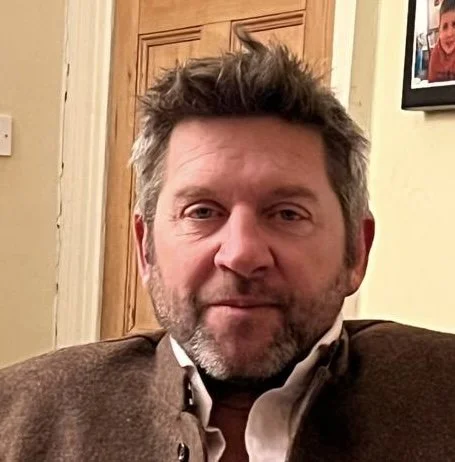 A middle-aged man with gray hair and a beard sitting in a room with wooden panels behind him.