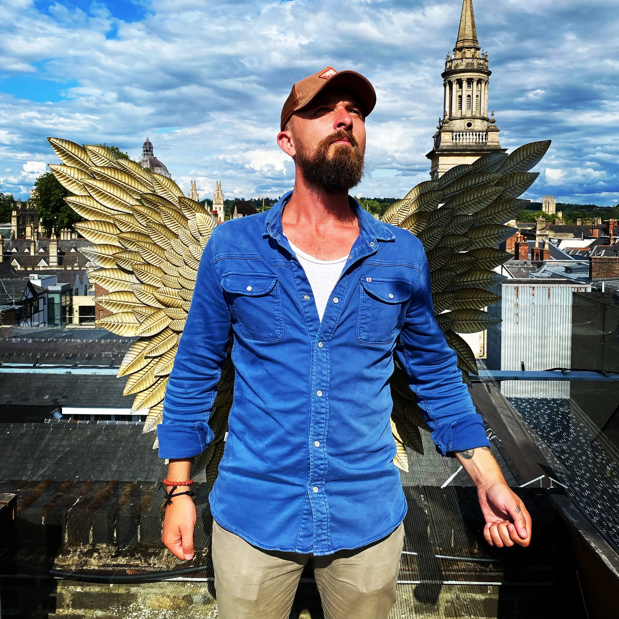 A man with a beard and mustache wearing a brown cap, blue jacket, white shirt, and beige pants stands on a rooftop with metallic angel wing decorations behind him. The background features historic buildings and a cloudy sky.