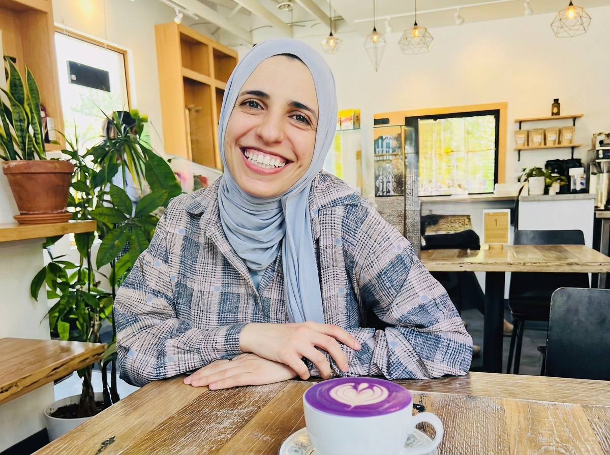 A woman, Sana, wearing a blue hijab smiles at a coffee shop table.