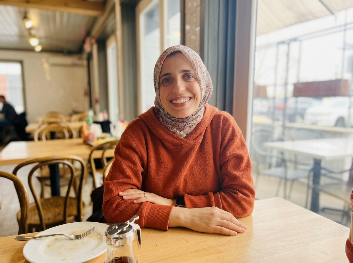 A woman, Sana Jaber, wearing a paisley hijab and an orange sweatshirt warmly smiles while sitting at a restaurant table.