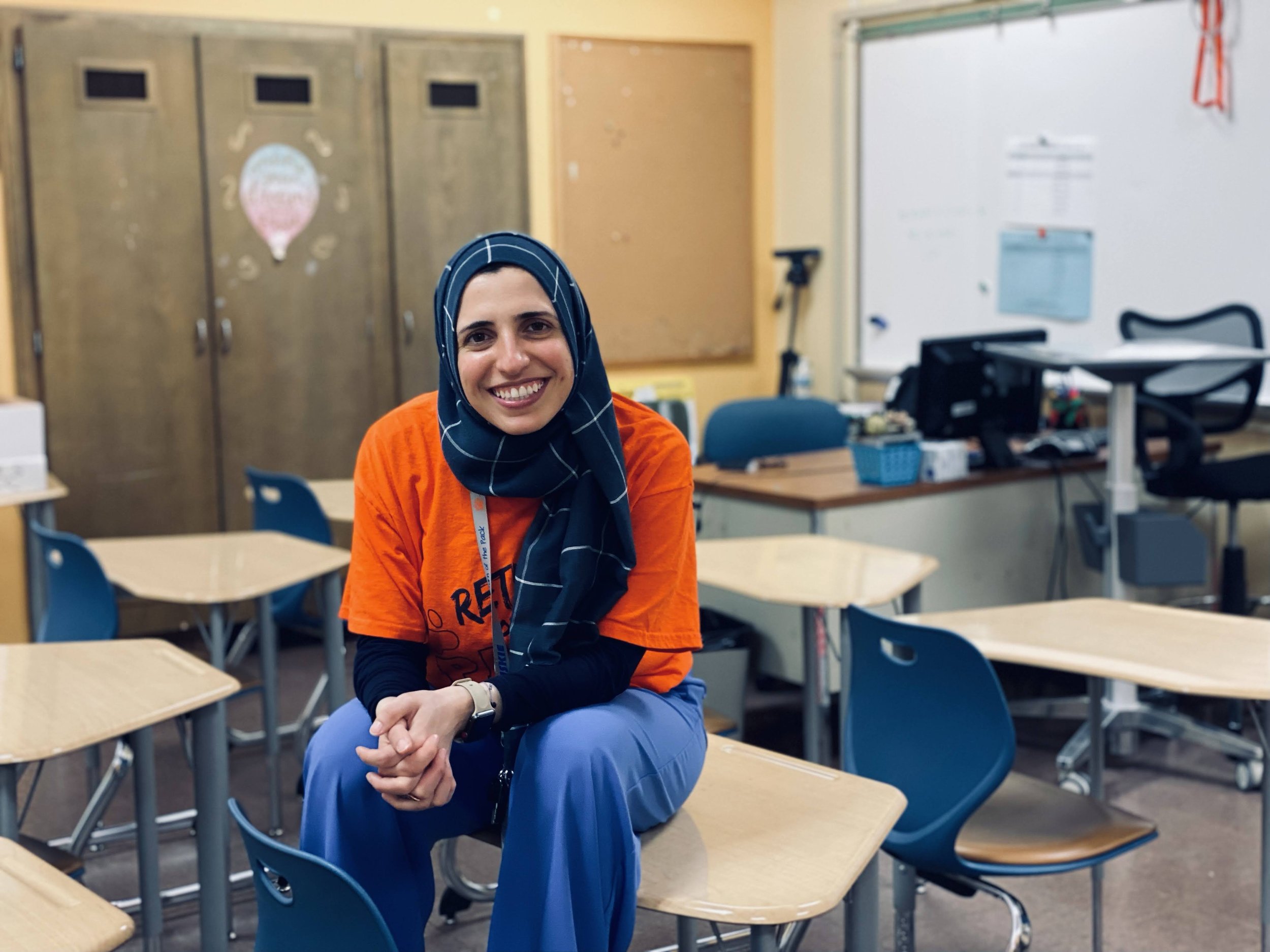 Teacher Sana Jaber sits on top of a desk and smiles in her high school math classroom.