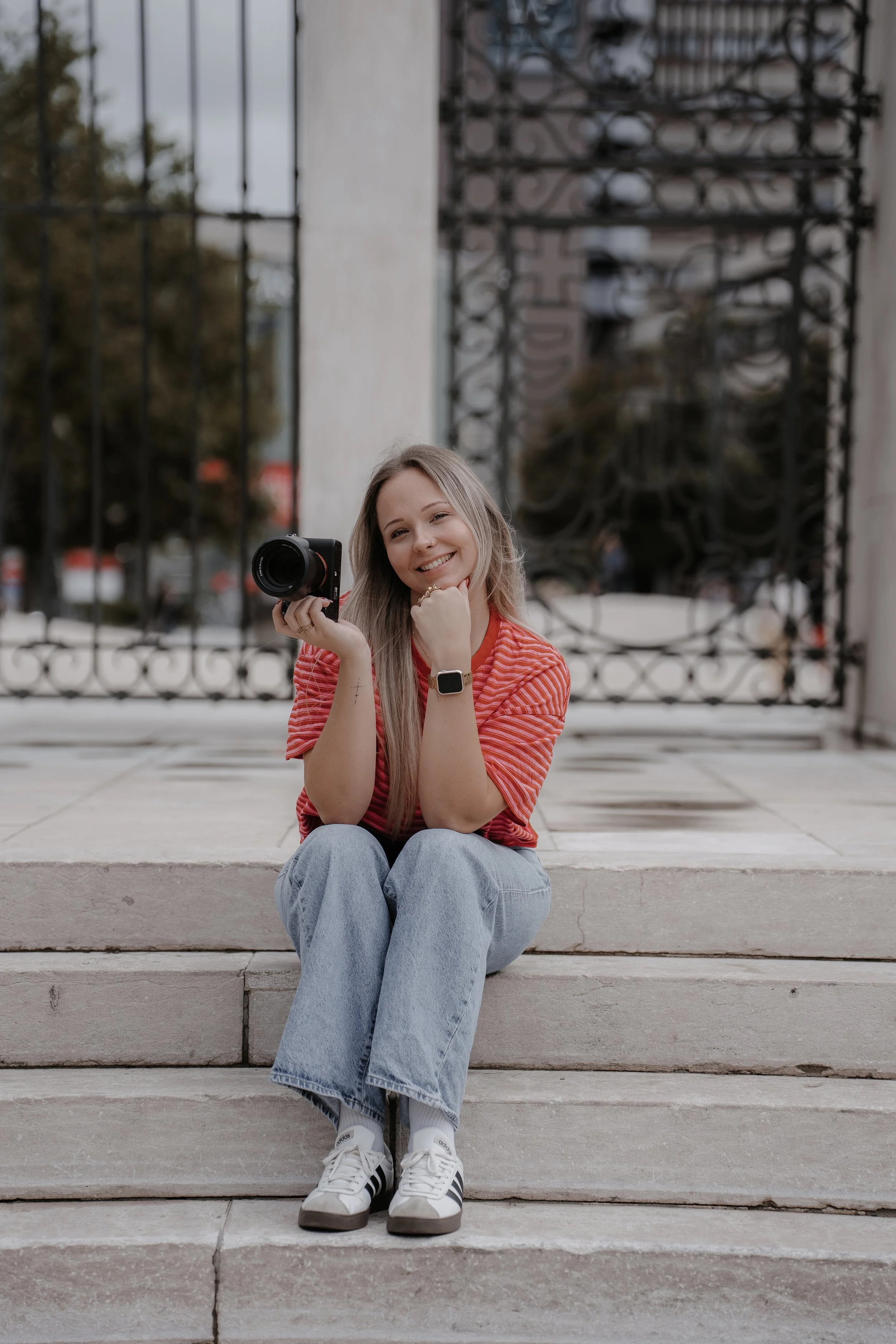 Junge Frau mit langen blonden Haaren, lächelnd, sitzt auf Steinstufen vor einem metallischen Tor, hält eine Kamera in der Hand, trägt ein rotes gestreiftes T-Shirt, eine Smartwatch und weiße Sportschuhe.