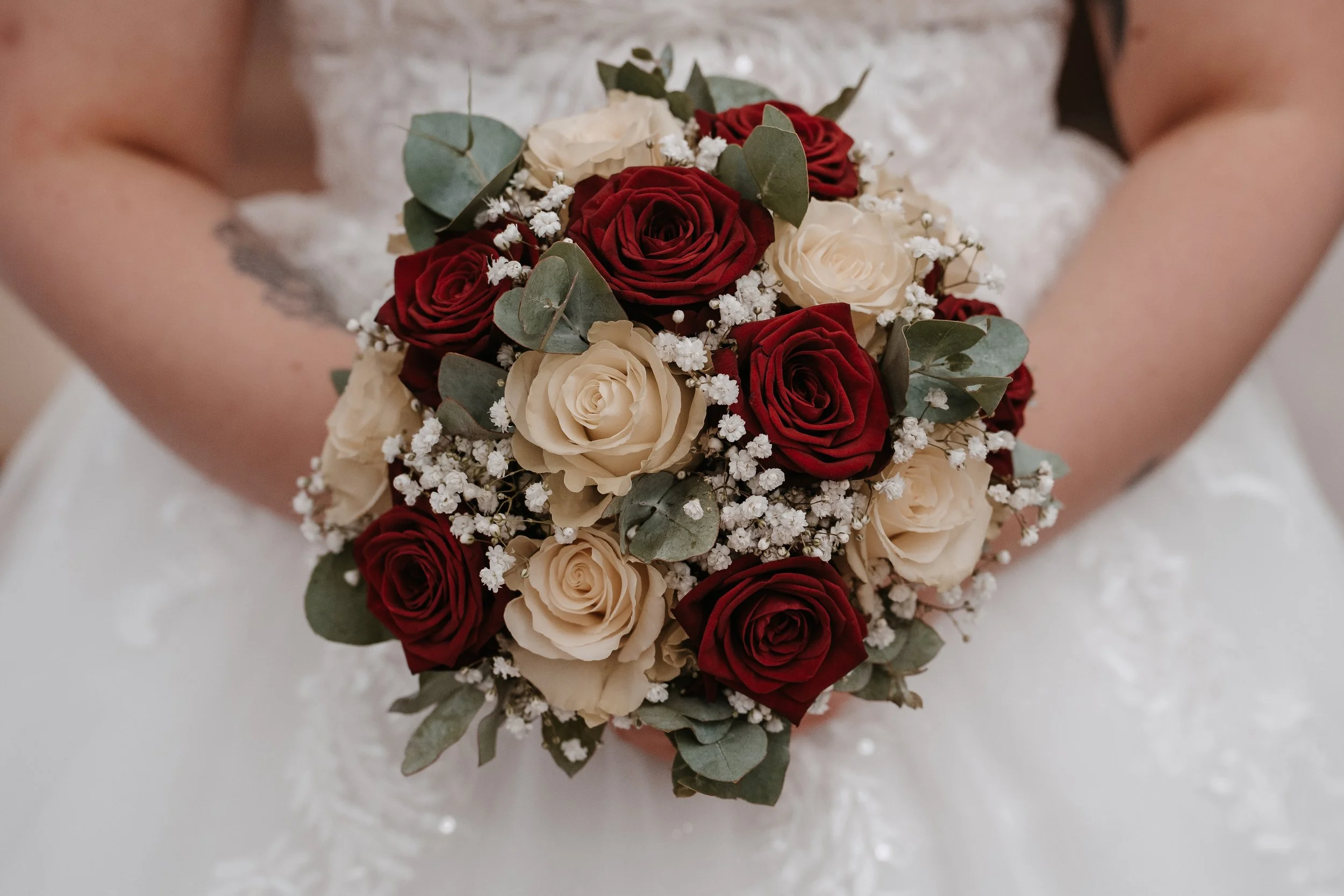 Ein Brautstrauß aus roten und weißen Rosen, begleitet von kleinen weißen Blüten und grünem Eukalyptus, gehalten von einer Person in einem weißen Hochzeitskleid.