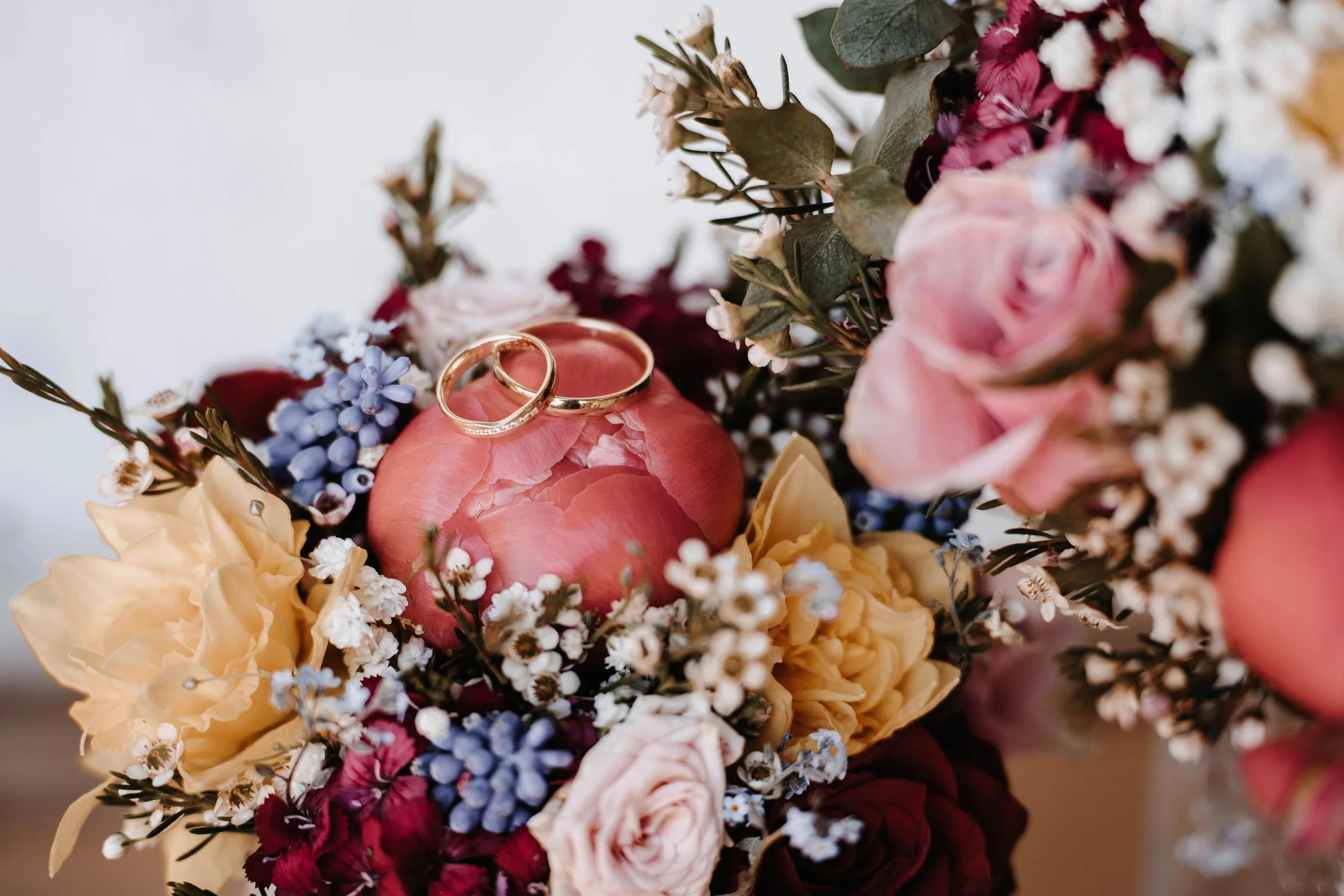 Ein Blumenstrauß mit Rosen, kleinen blauen und weißen Blumen sowie grünen Blättern, mit zwei goldenen Eheringen auf einer rosa Entenblüte.