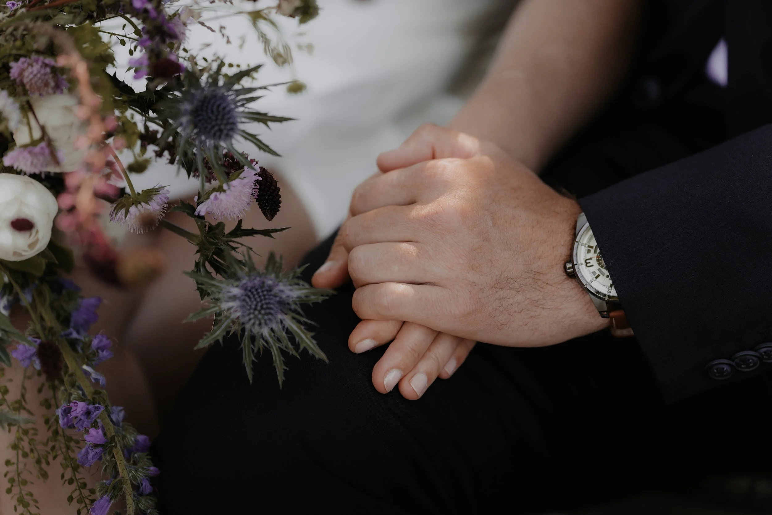 Nahaufnahme von zwei Händen, die aufeinander liegen, neben einem Blumenstrauß mit lila, weißen und dunkelvioletten Blumen. Jemand trägt ein schwarzes Sakko und eine Uhr am Handgelenk.