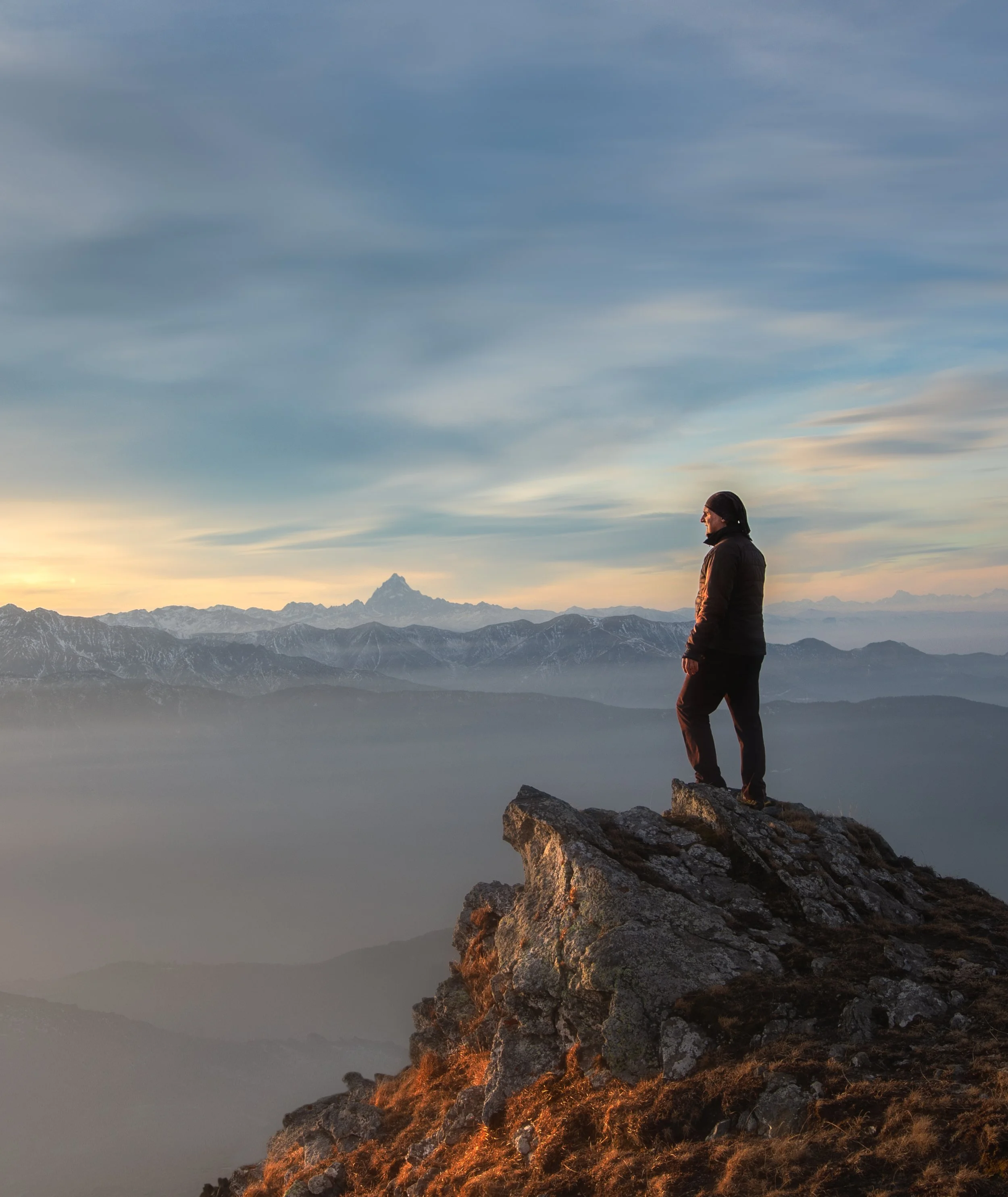 A person standing on a rocky ledge overlooking mountain ranges at sunset or sunrise, with a mostly cloudy sky and a distant mountain peak.