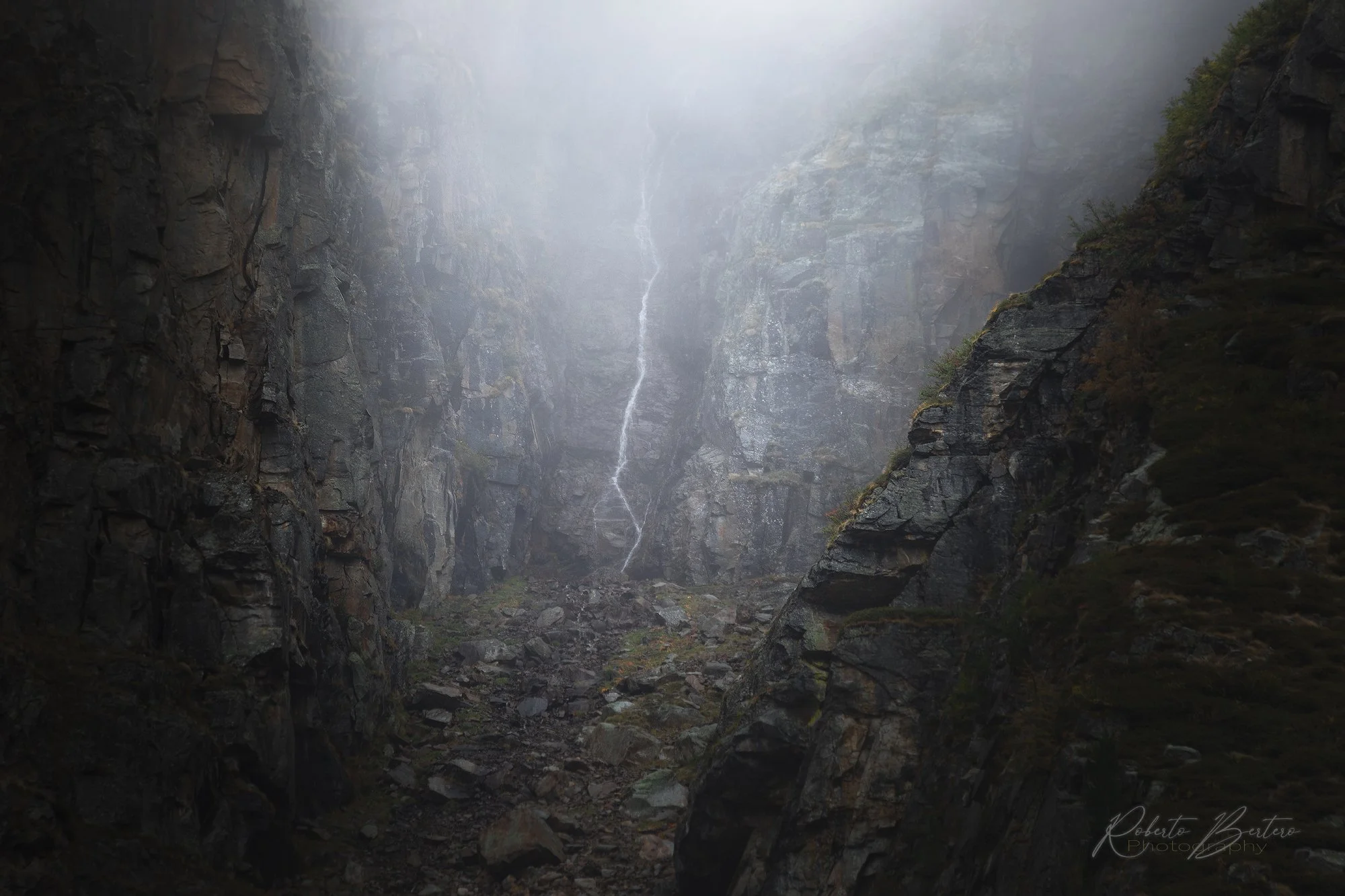 A foggy mountain gorge with steep rocky walls on either side and a narrow stream of water descending from the top, with mist obscuring the upper part of the scene.