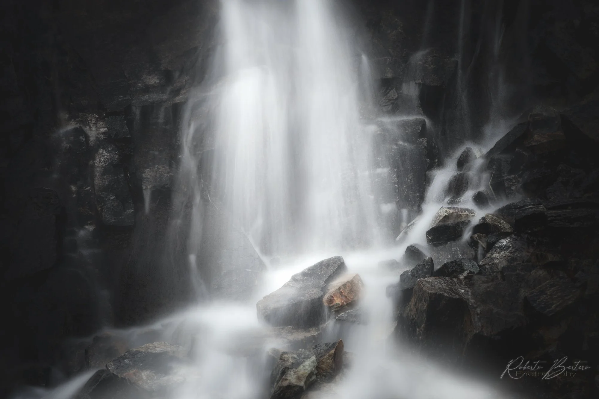 A waterfall cascading down dark rocks in a dimly lit environment, with white water flowing over the rocks and splashing at the bottom.